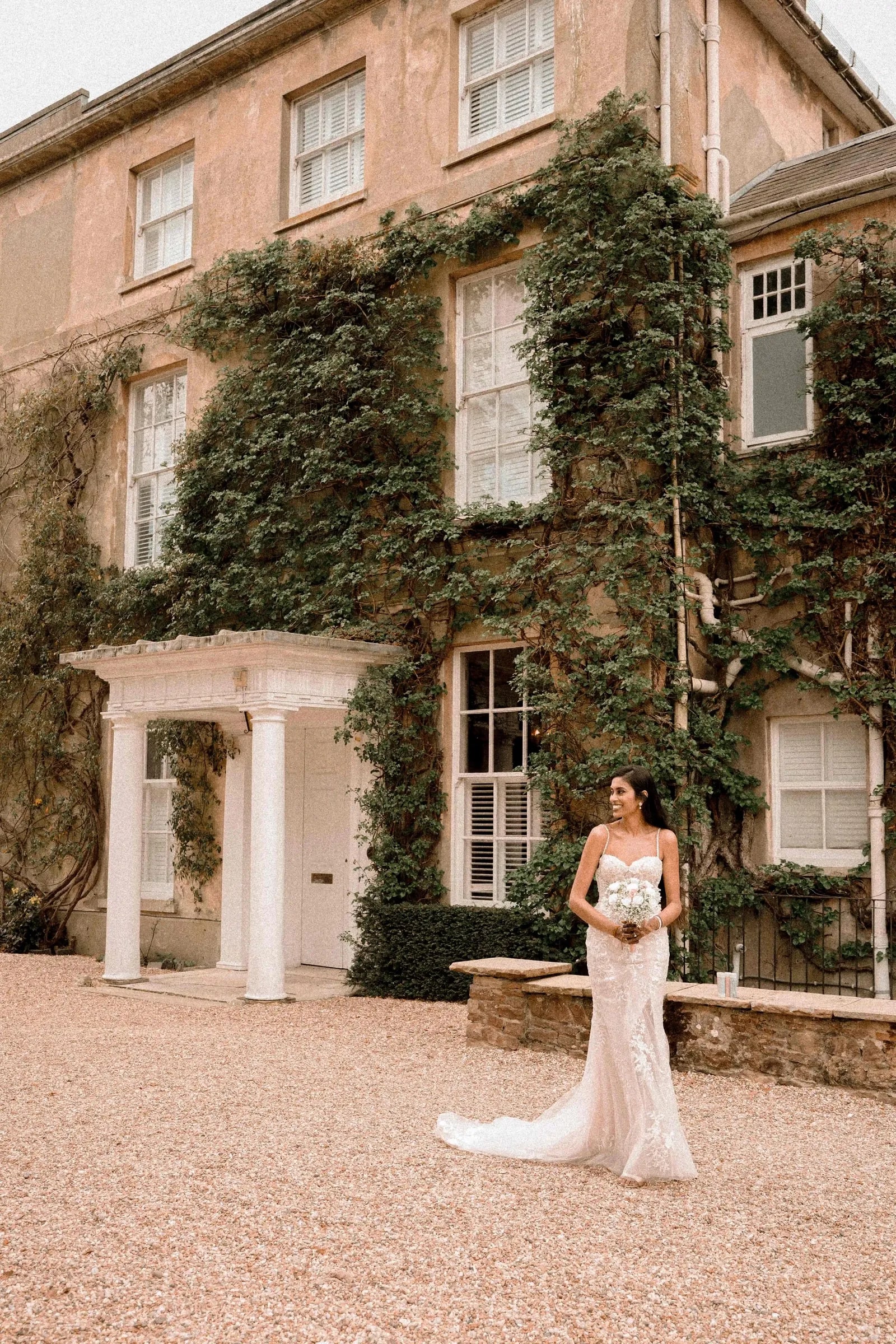 Bride alone with her bouquet on the drive before the ivy-covered house