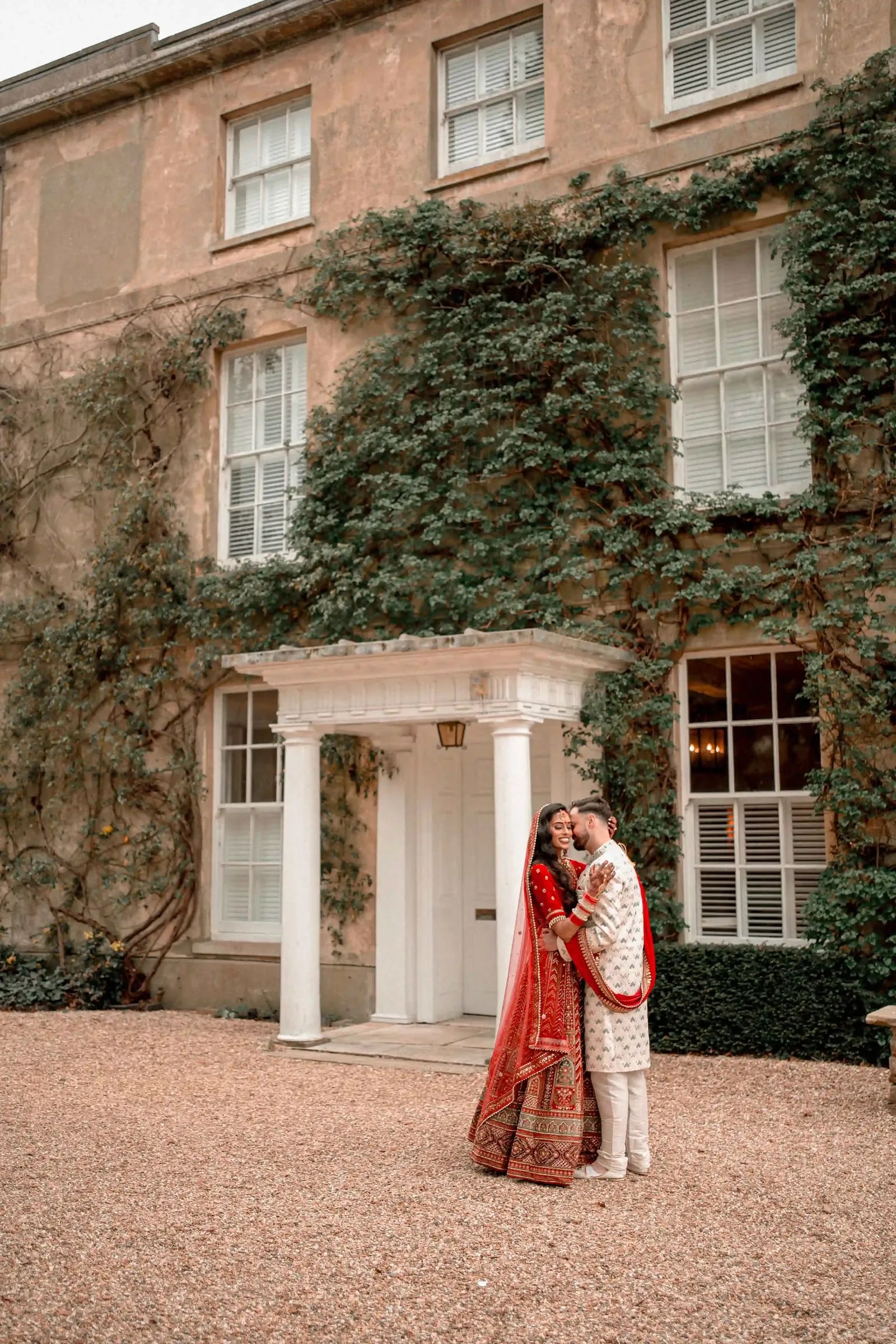Couple in red and ivory embracing under the portico of the ivy-covered house