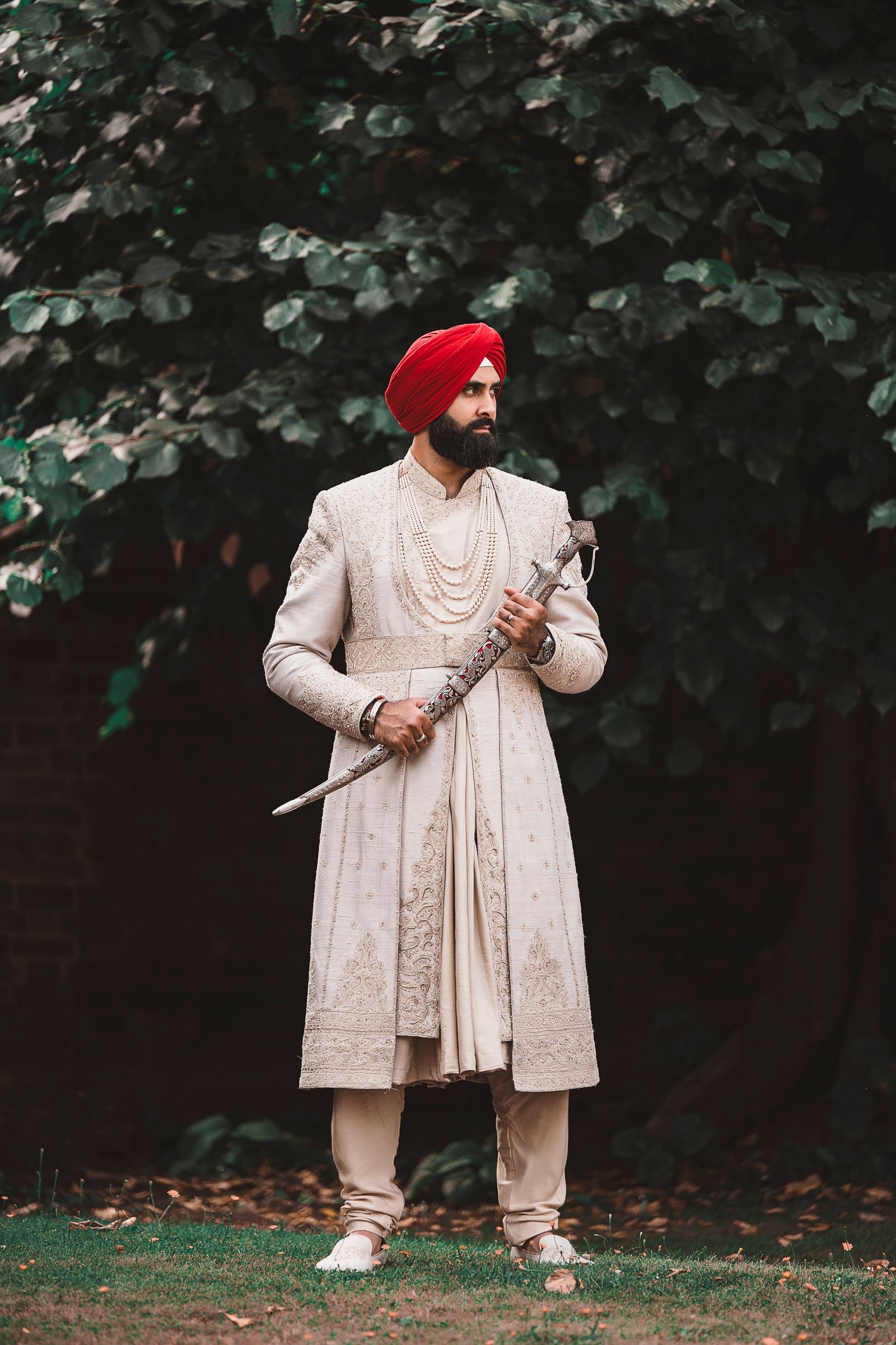 Sikh groom in a red turban and ivory sherwani holding his kirpan, standing before a dark copper beech