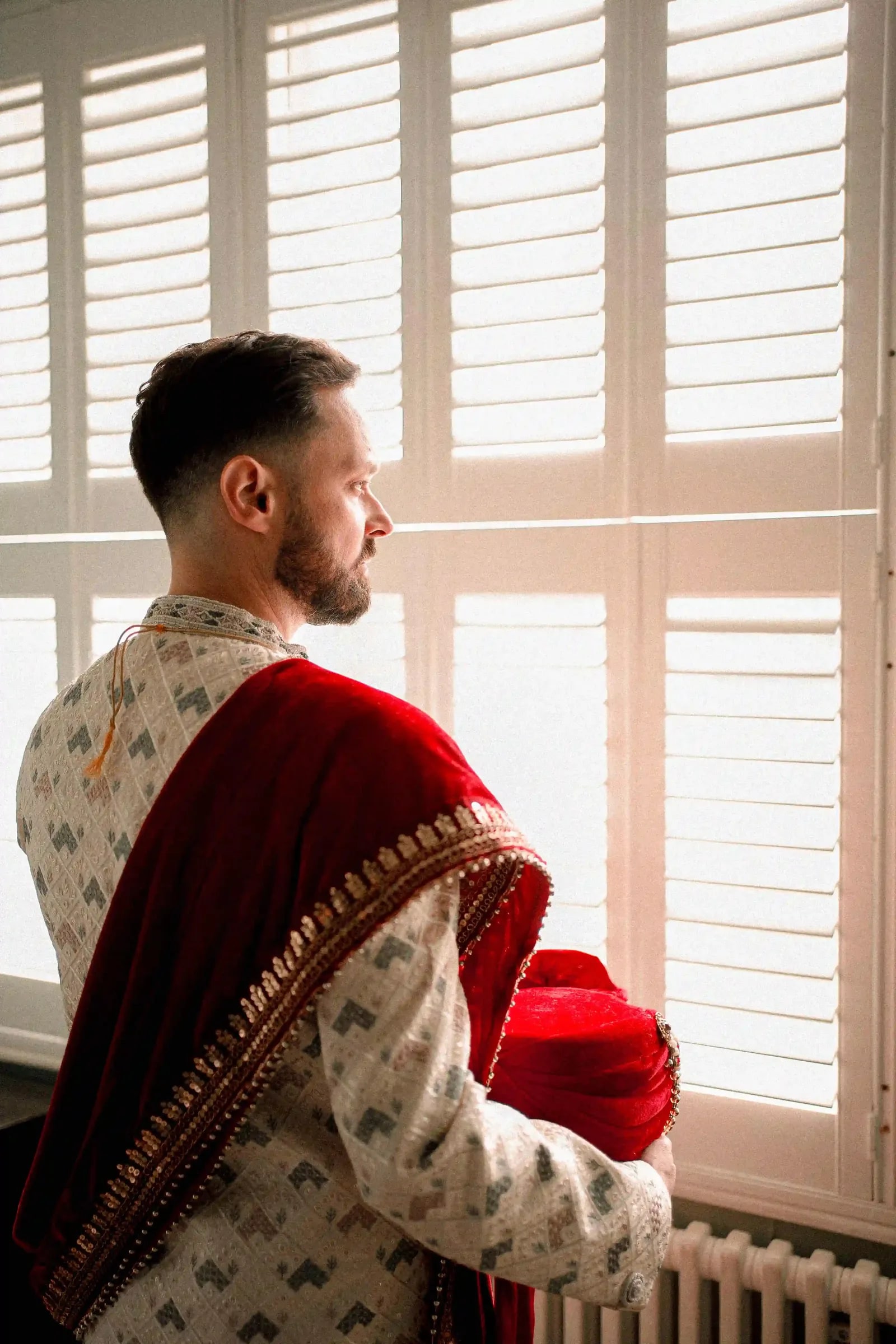 Groom in an ivory sherwani and red shawl holding his turban at the window