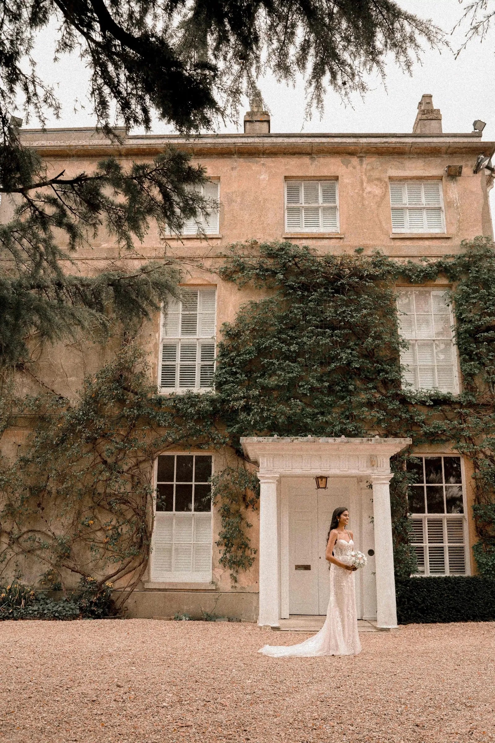 Bride standing small in the doorway of the house, cedar branches overhead