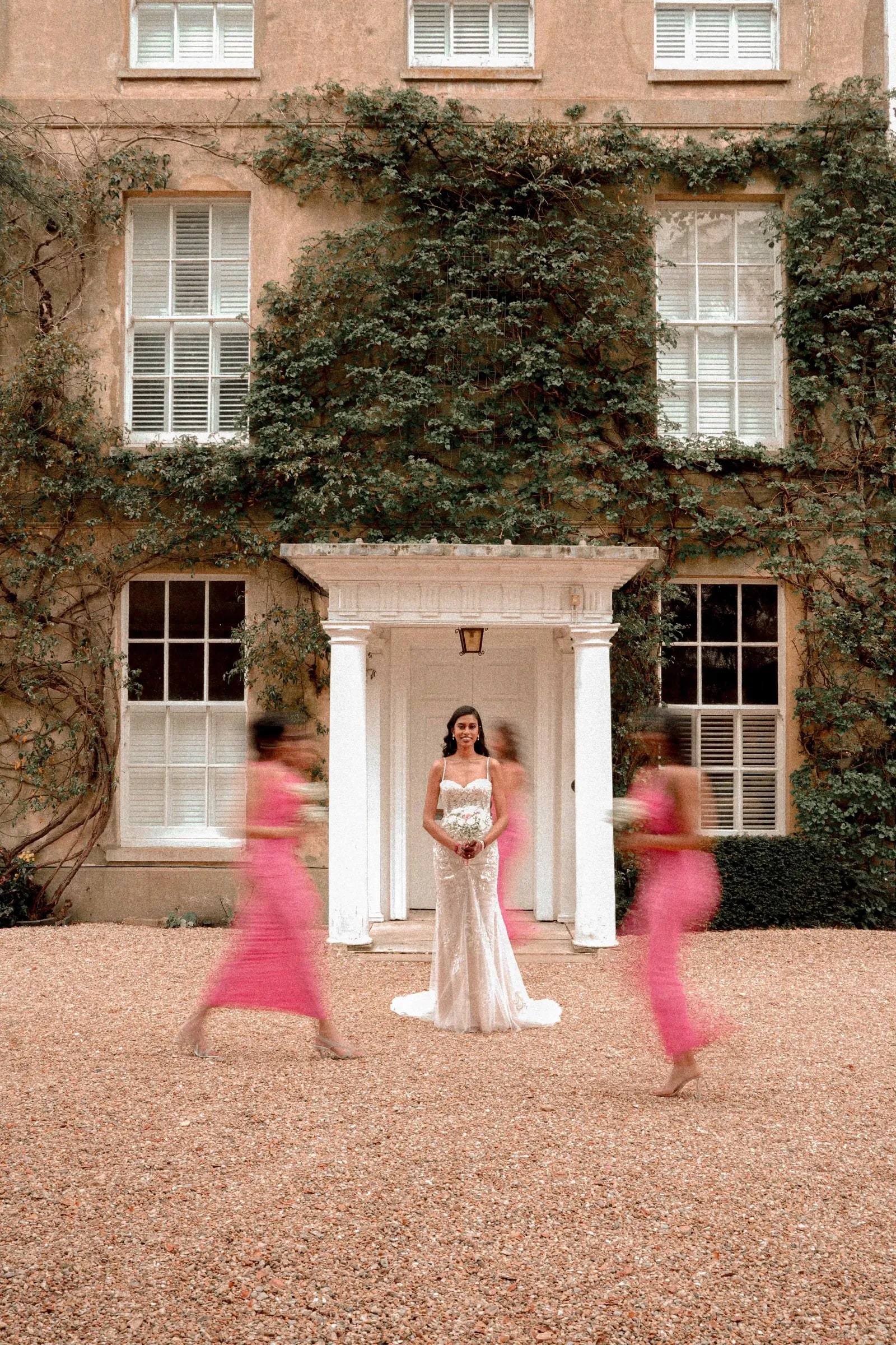 Bride still in the doorway as her bridesmaids in pink rush past, blurred with movement
