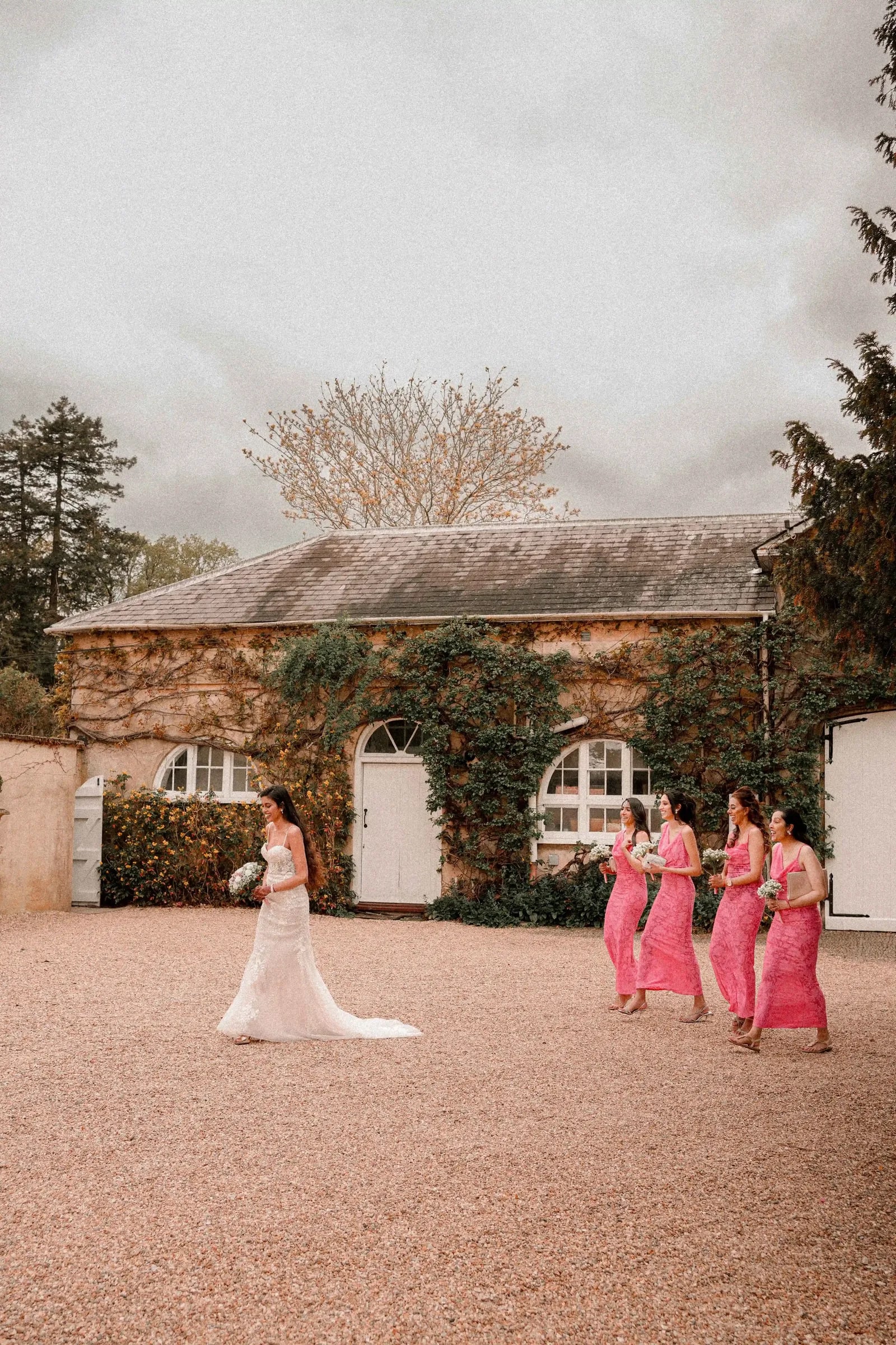 Bride at a distance on the gravel courtyard, her bridesmaids waiting to one side
