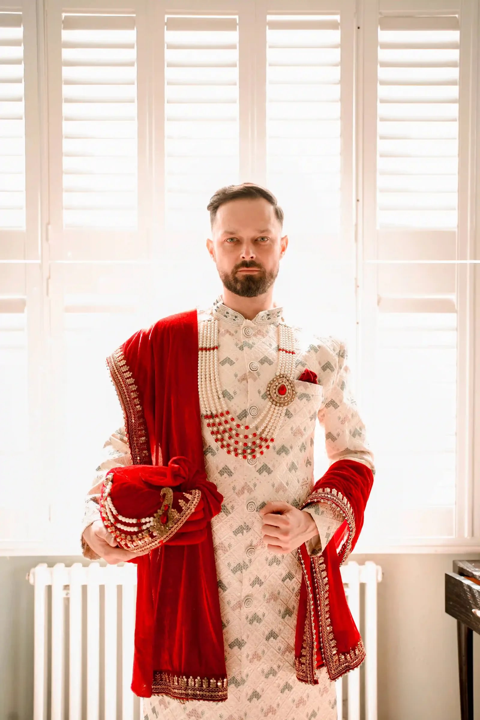 Groom standing at the shuttered window in an ivory sherwani with a red velvet shawl