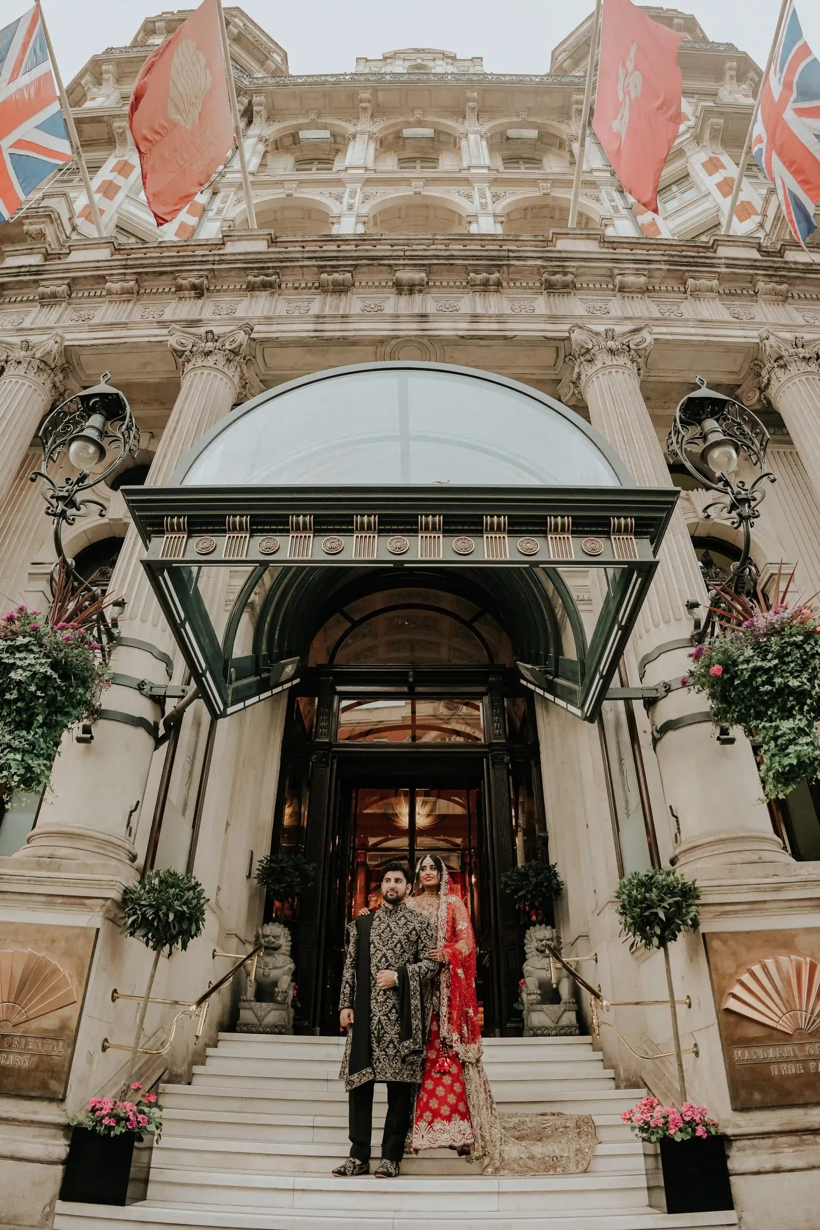 Couple small beneath the glass canopy of the hotel entrance, flags along the facade