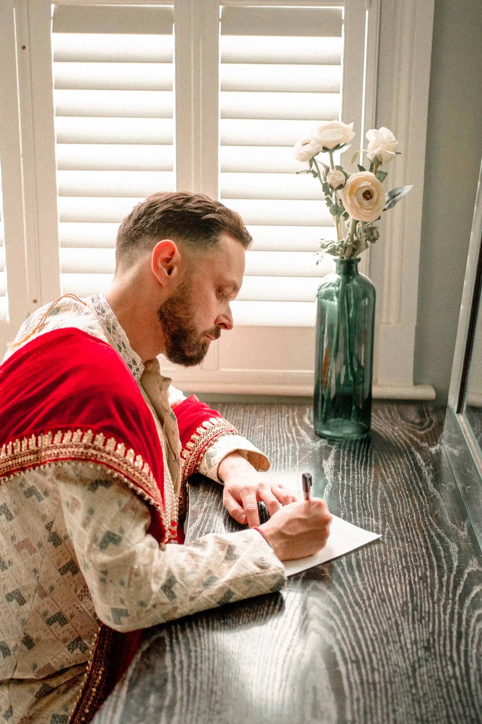 Groom writing a note at a table, a vase of white ranunculus beside him