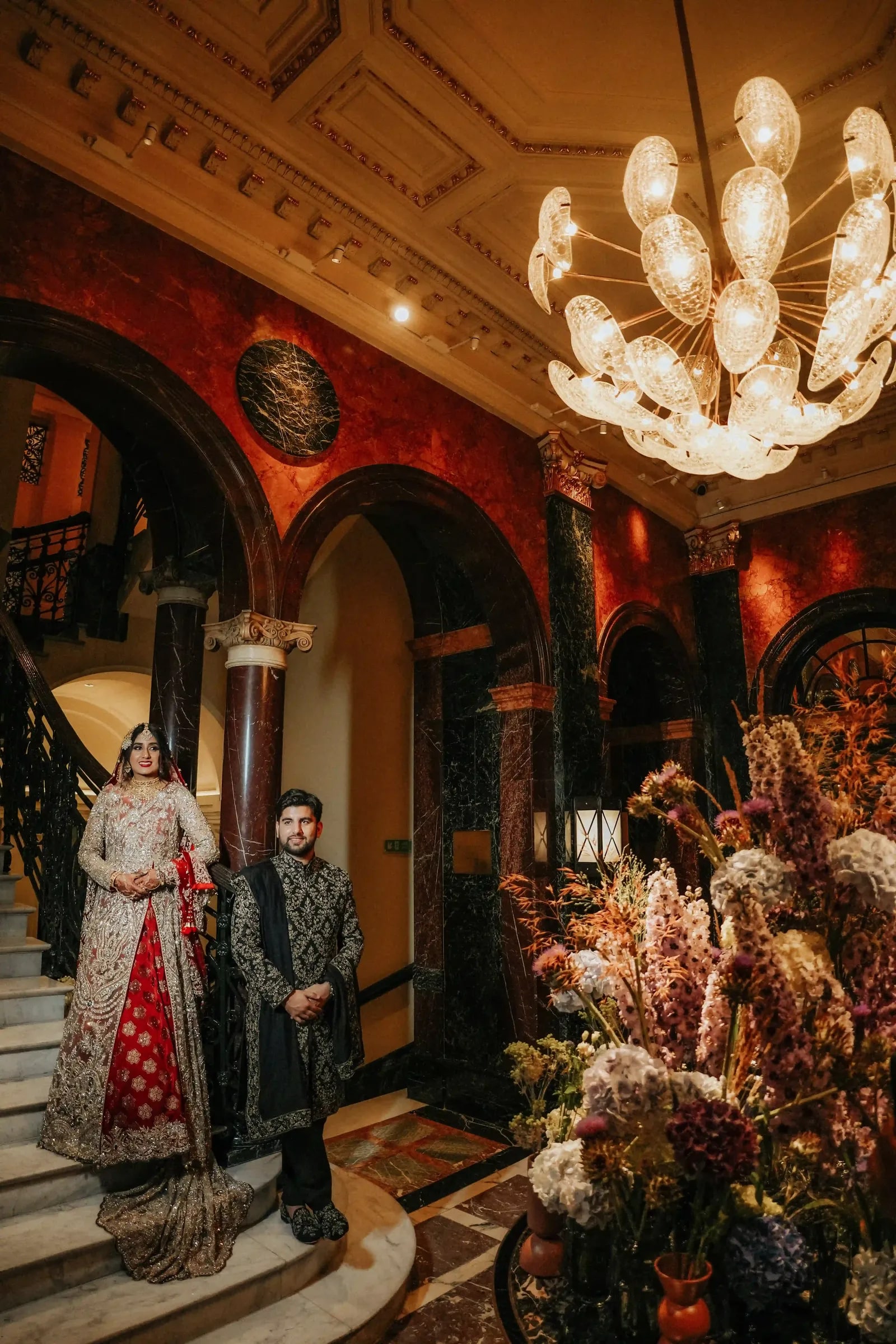 Couple in the hotel lobby beneath a chandelier, banked flowers beside them