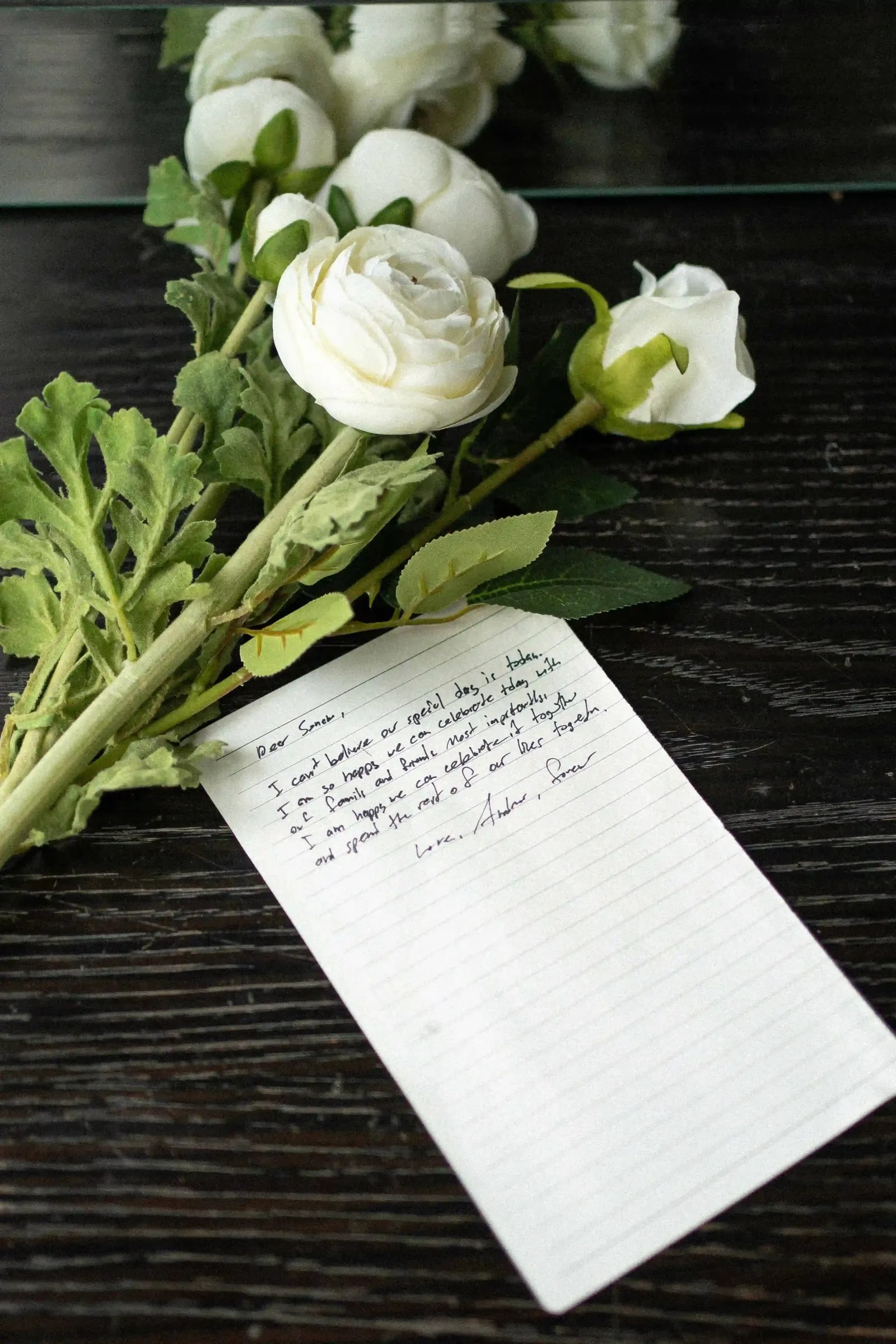 A handwritten letter and white ranunculus laid on a dark wooden table