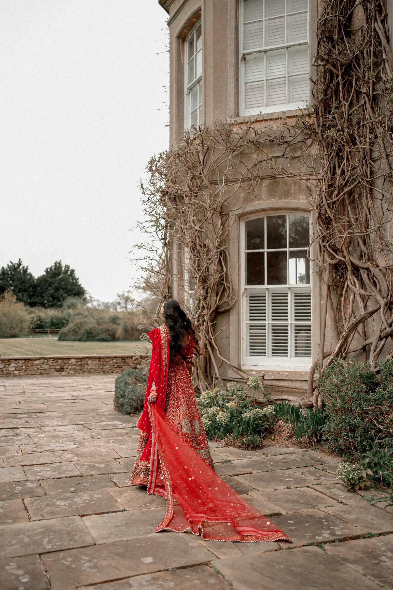 Bride walking the terrace of the ivy-covered house, her red dupatta trailing