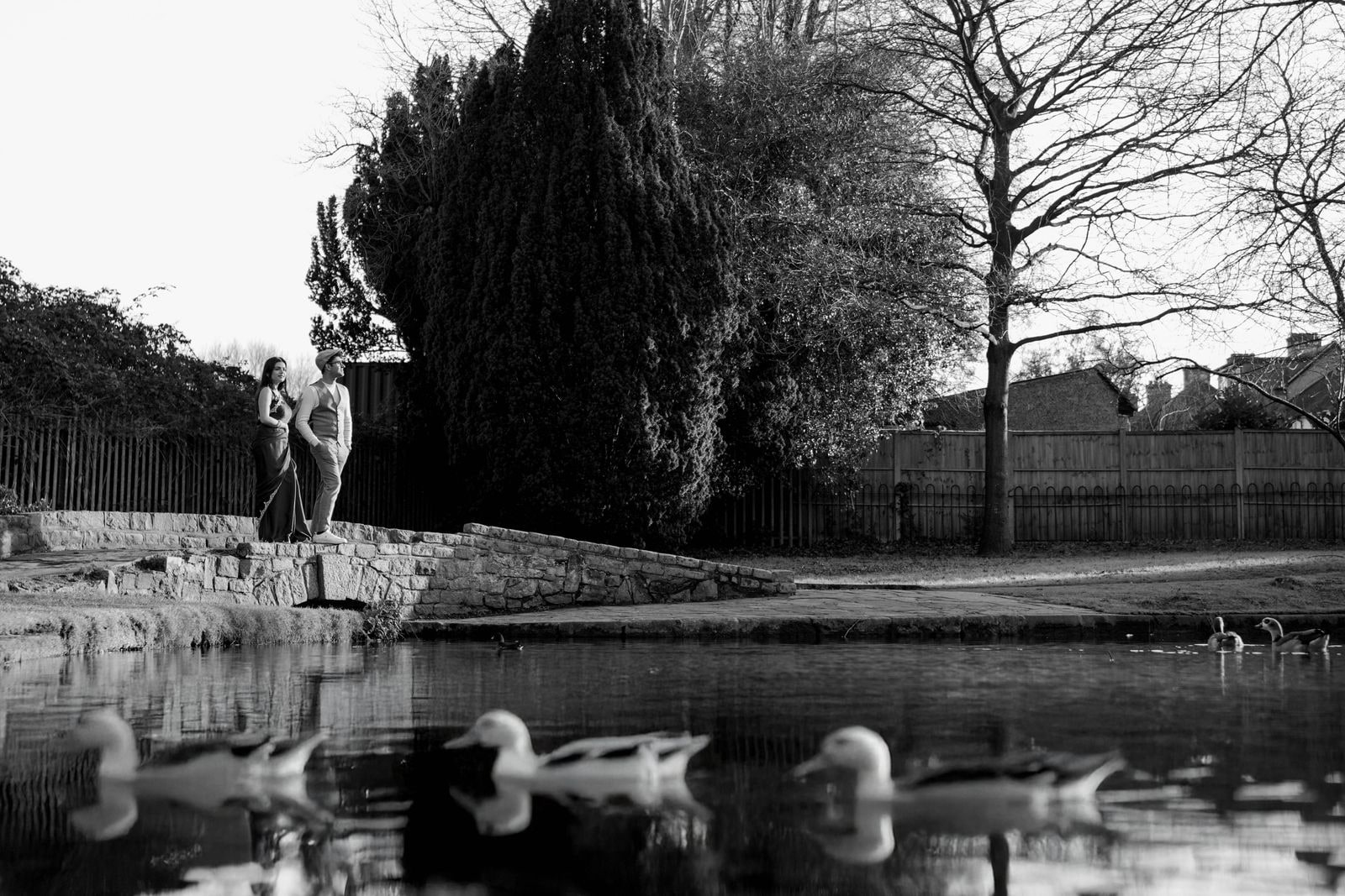 A figure by the pond with ducks on the water, bare trees behind, in black and white