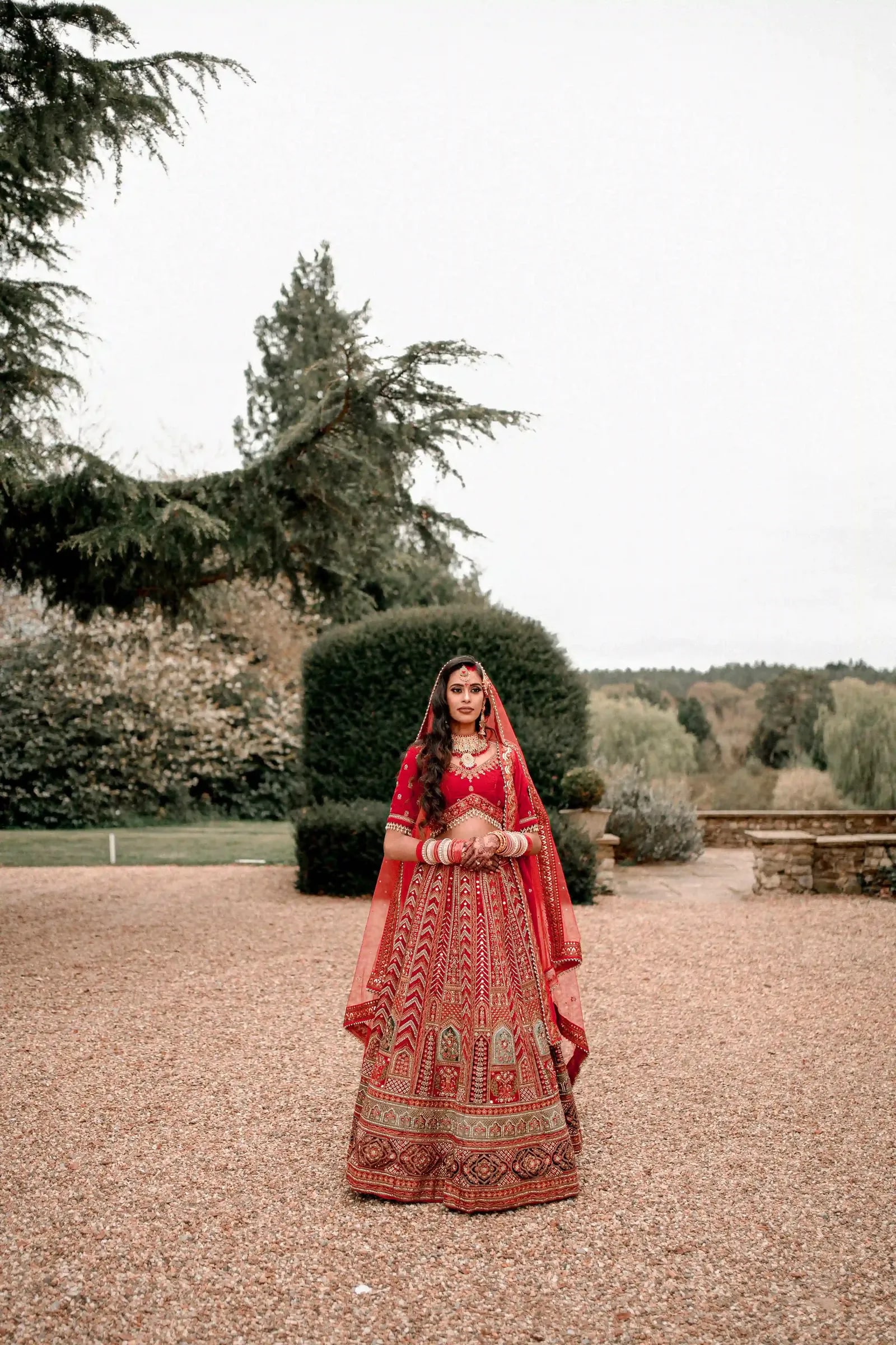 Bride alone on the gravel drive in a red and gold lehenga, parkland behind her