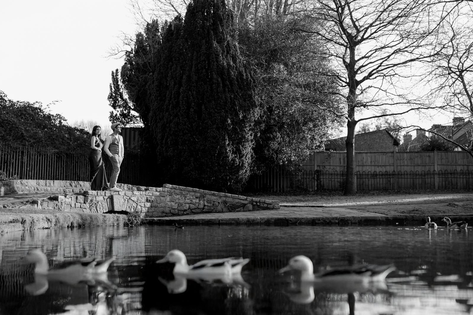 Figure standing at the edge of a pond with ducks on the water, in black and white