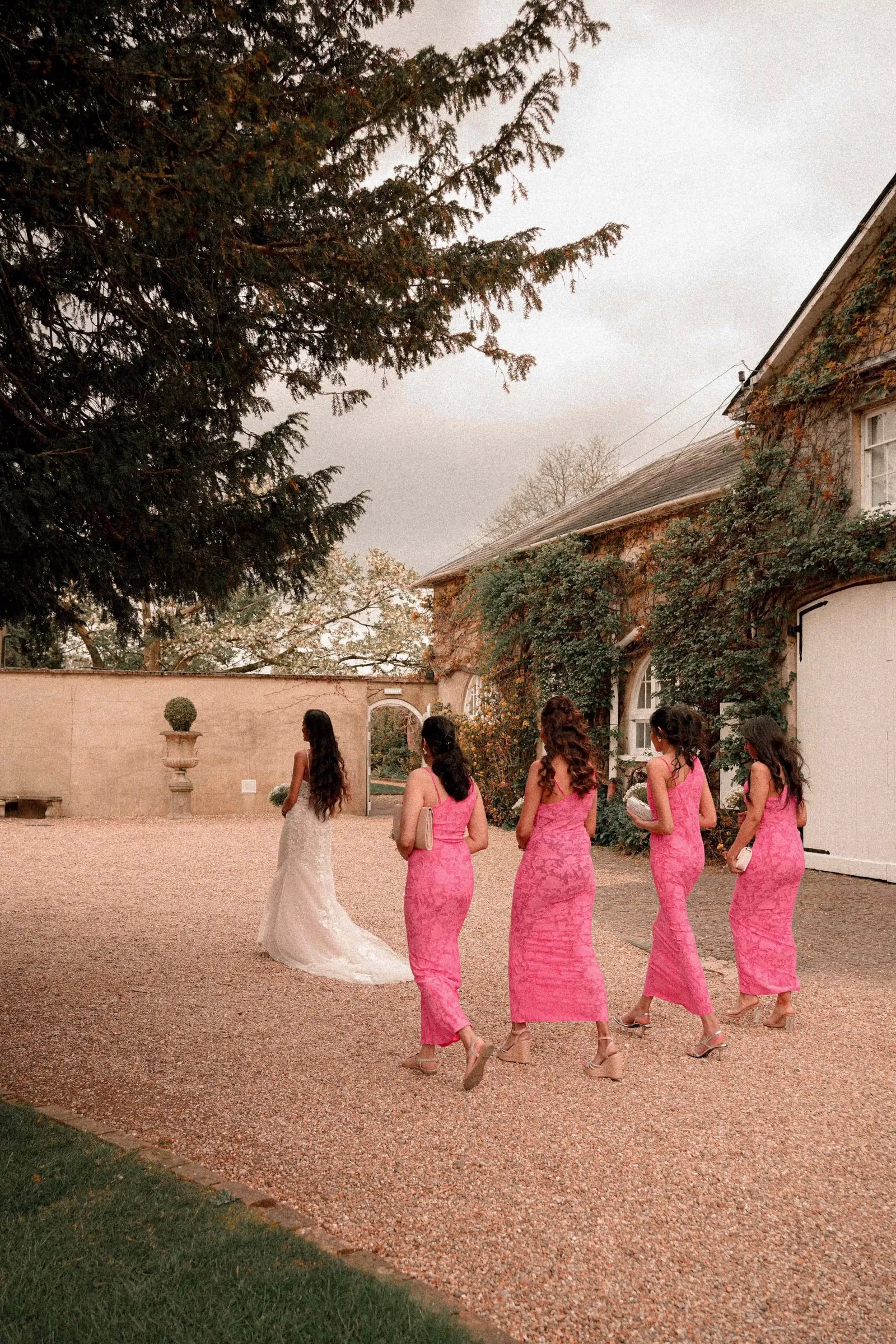 Bridesmaids in pink walking the bride along the drive towards the house