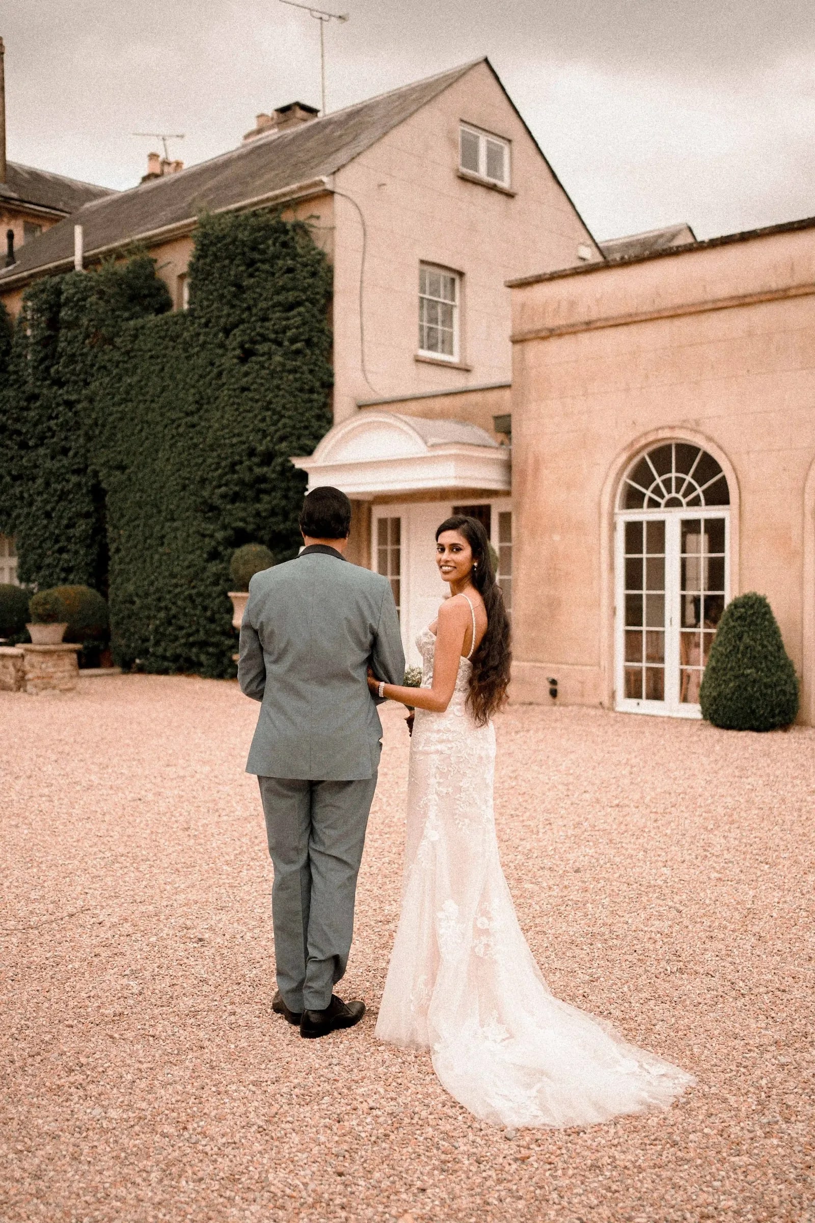 Bride and groom seen from behind on the gravel, facing the house