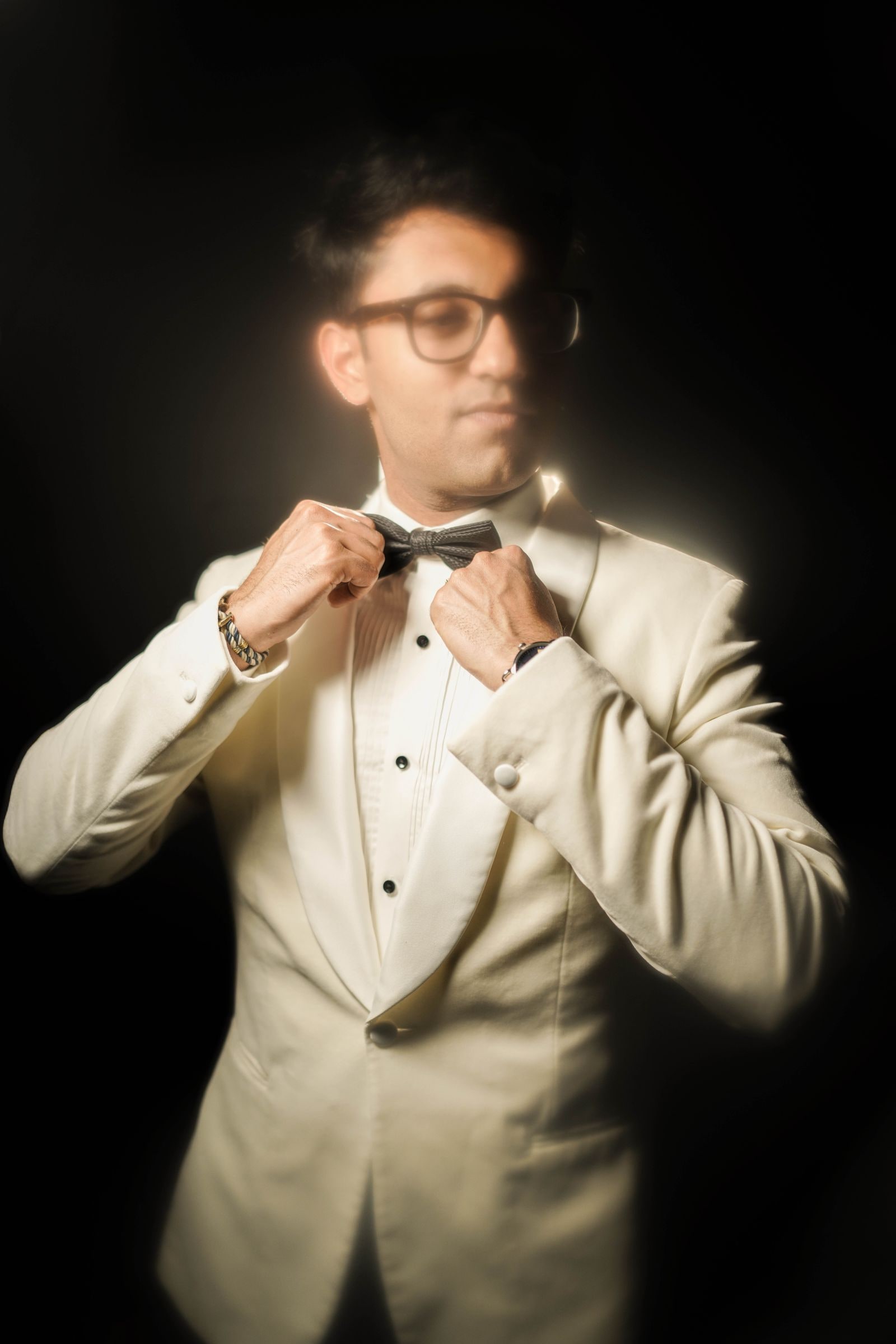 Groom adjusting his bow tie in a cream dinner jacket, lit against black
