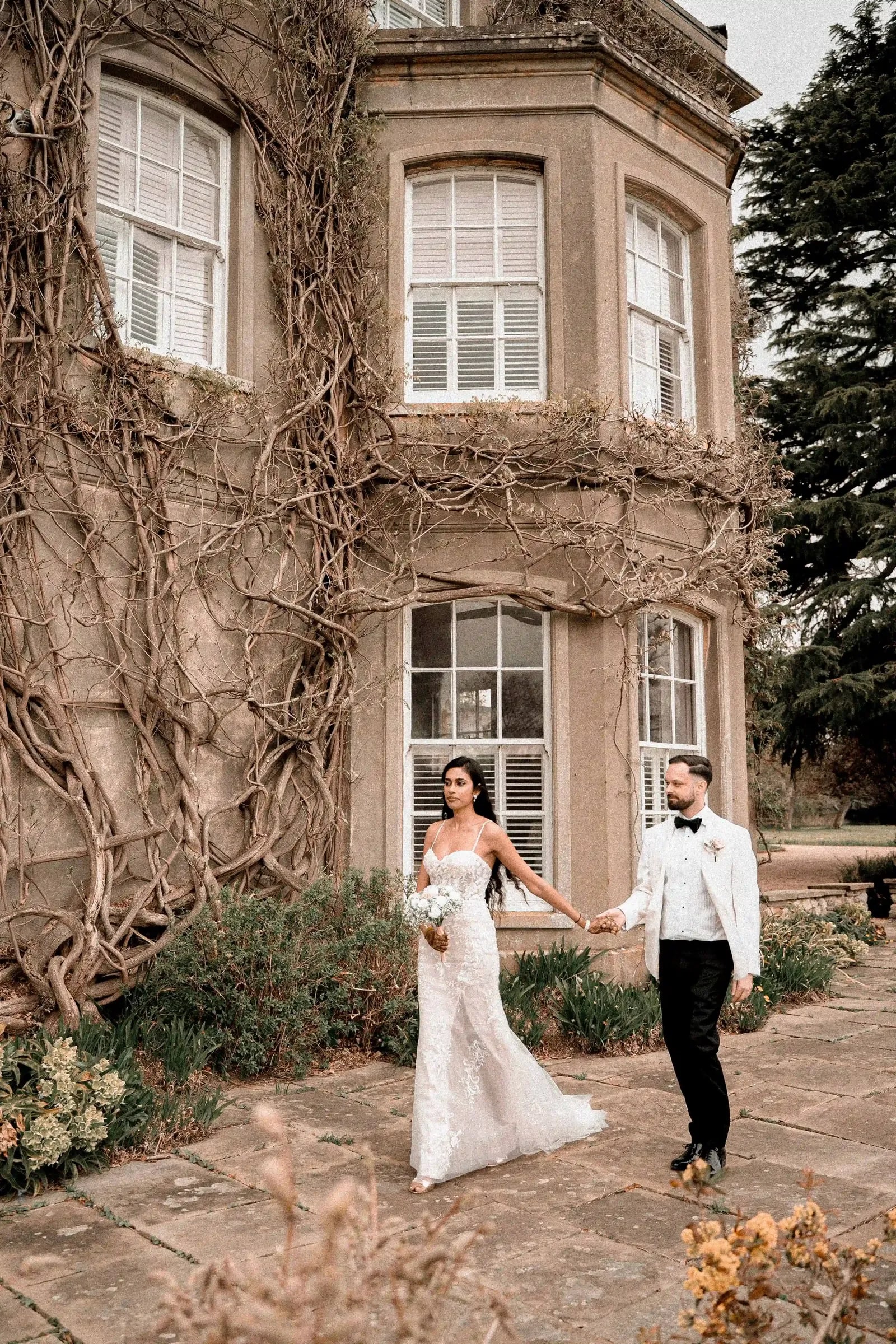 Bride in white and groom in a white dinner jacket holding hands beside the ivy-covered wall