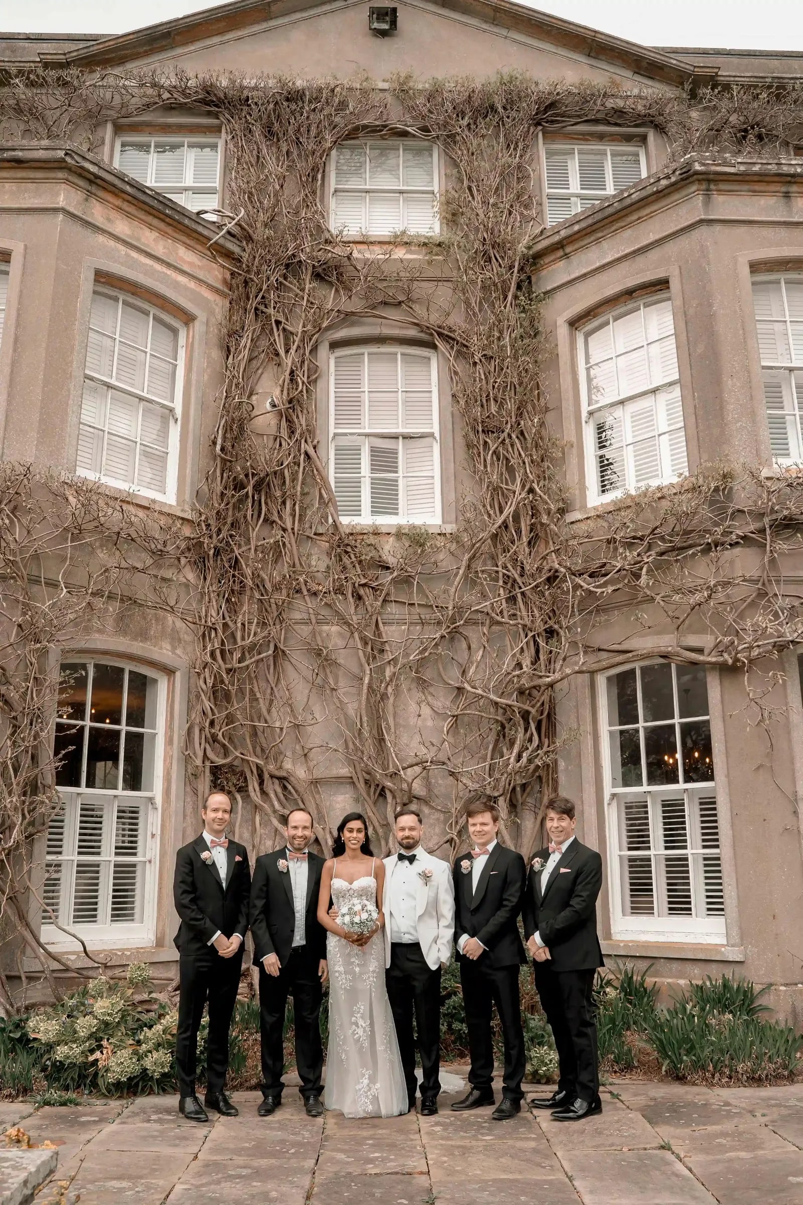 Bride and groom with the groomsmen in black tie before the ivy-covered house