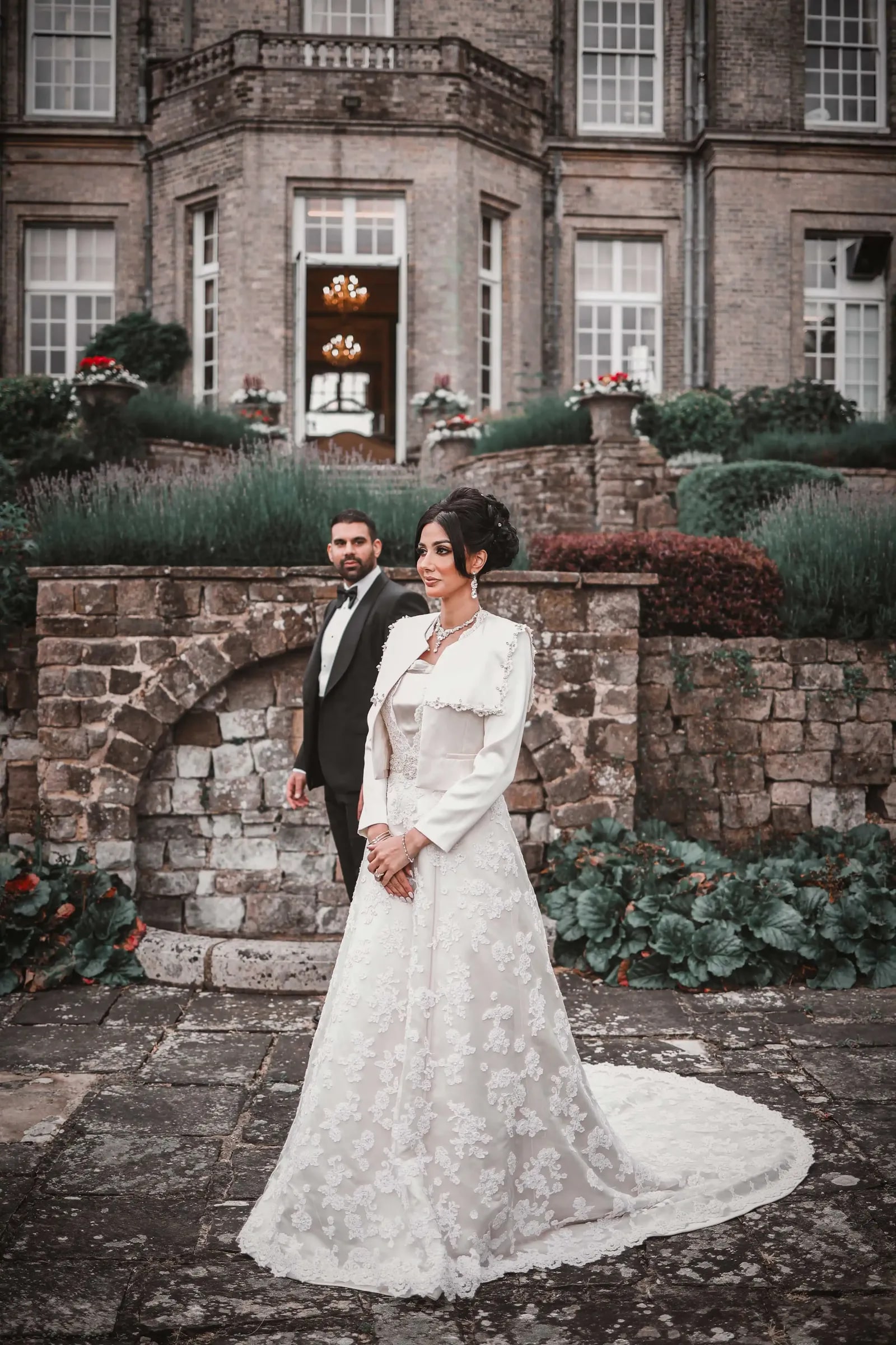 Bride in a lace gown on the terrace, her groom standing behind her against the stonework