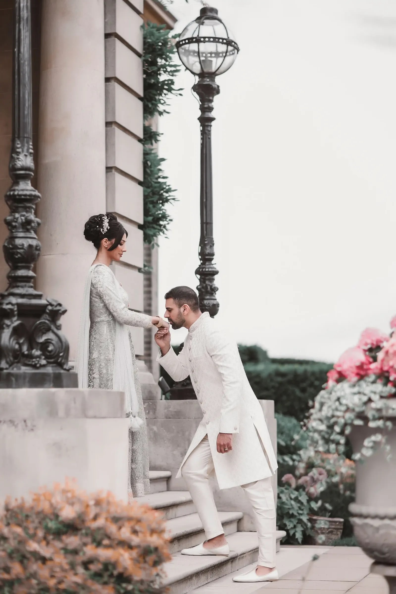 Groom in an ivory sherwani kneeling to kiss his bride's hand on the steps