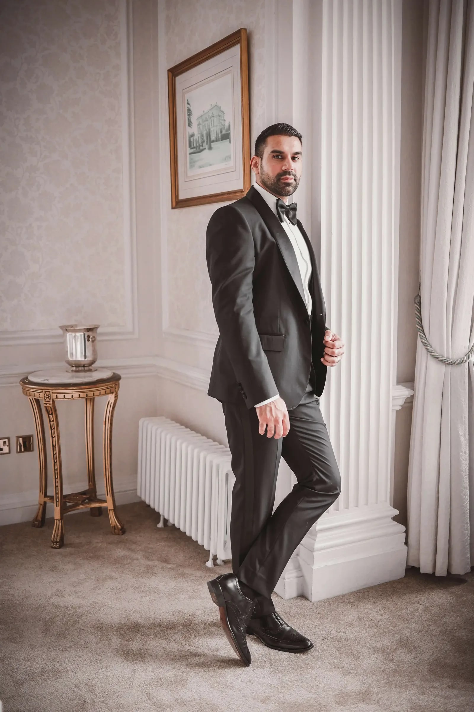 Groom in black tie standing by the window in a pale panelled room