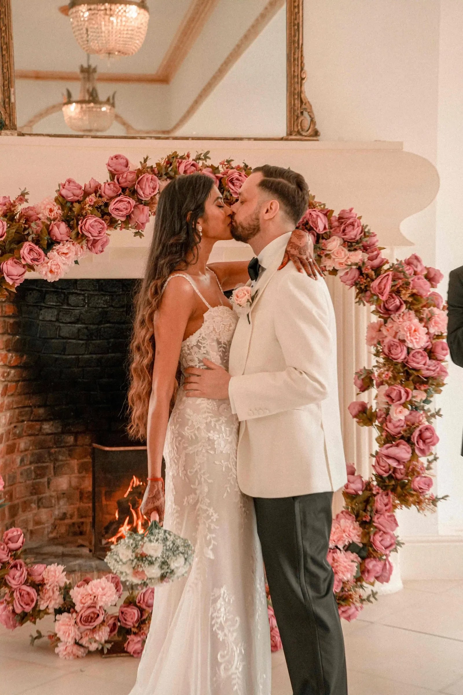 Bride and groom kissing before a pink rose arch and a lit fireplace