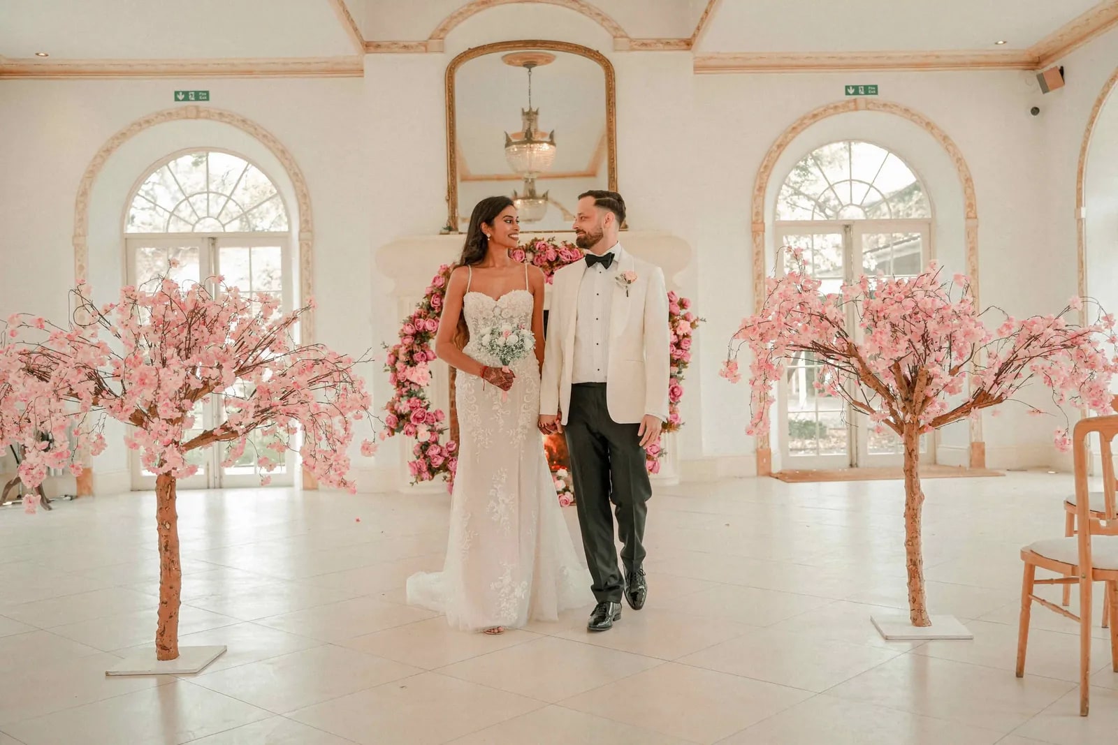 Bride and groom walking together through the pale ballroom between blossom trees