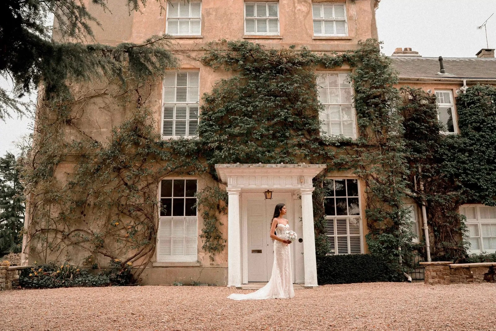 Bride alone in the doorway of the ivy-covered house, the full front in view