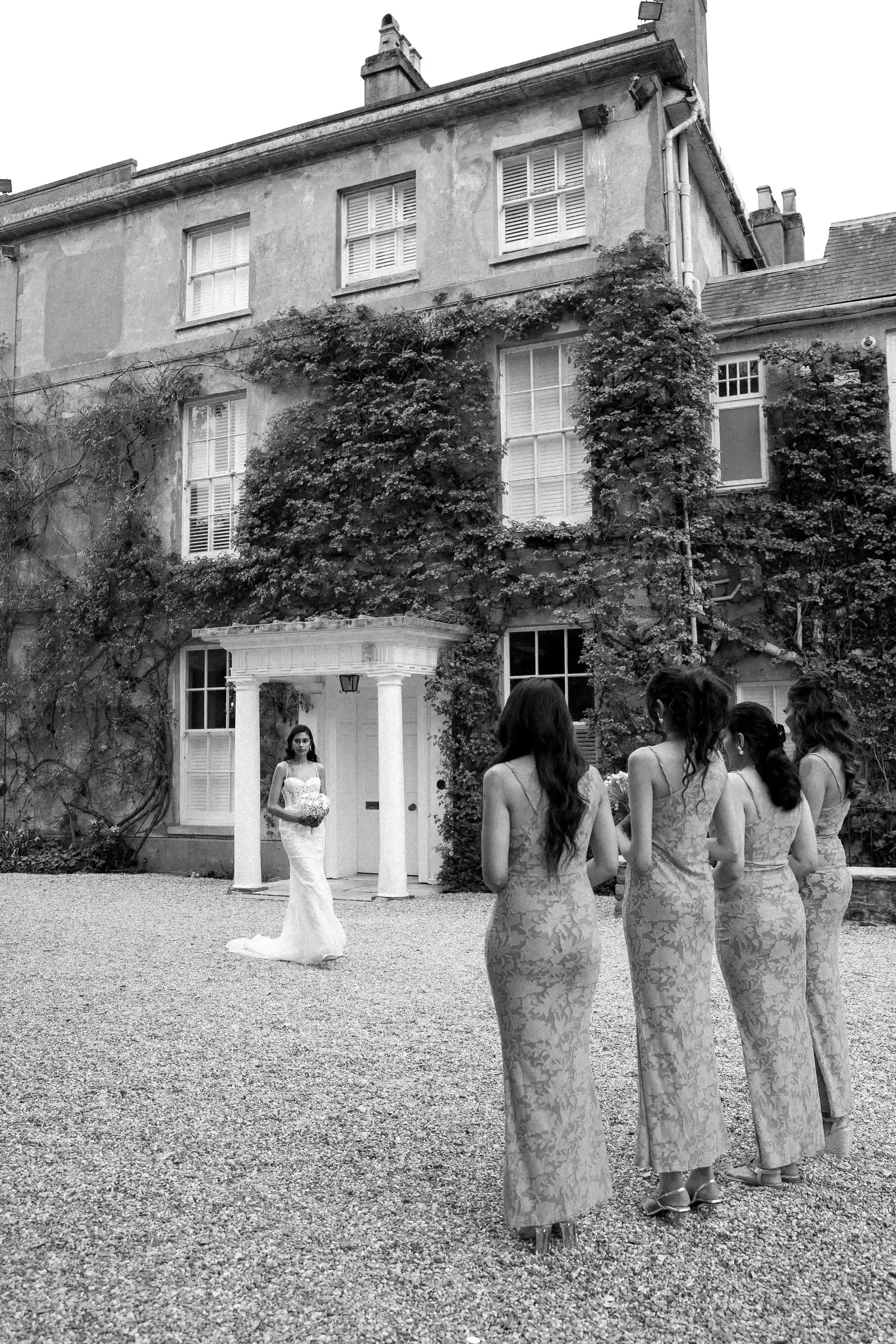 Bride waiting in the doorway as her bridesmaids walk towards her, in black and white