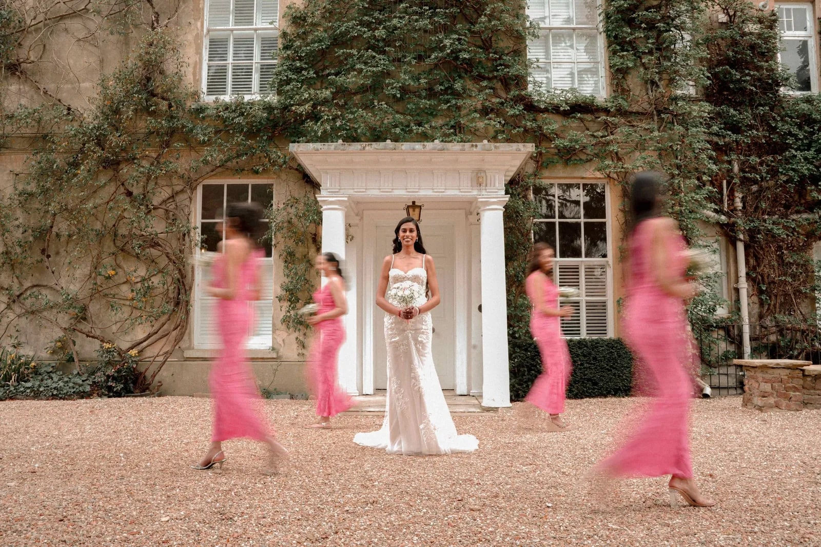 Bride still in the doorway while her bridesmaids sweep past in pink, blurred