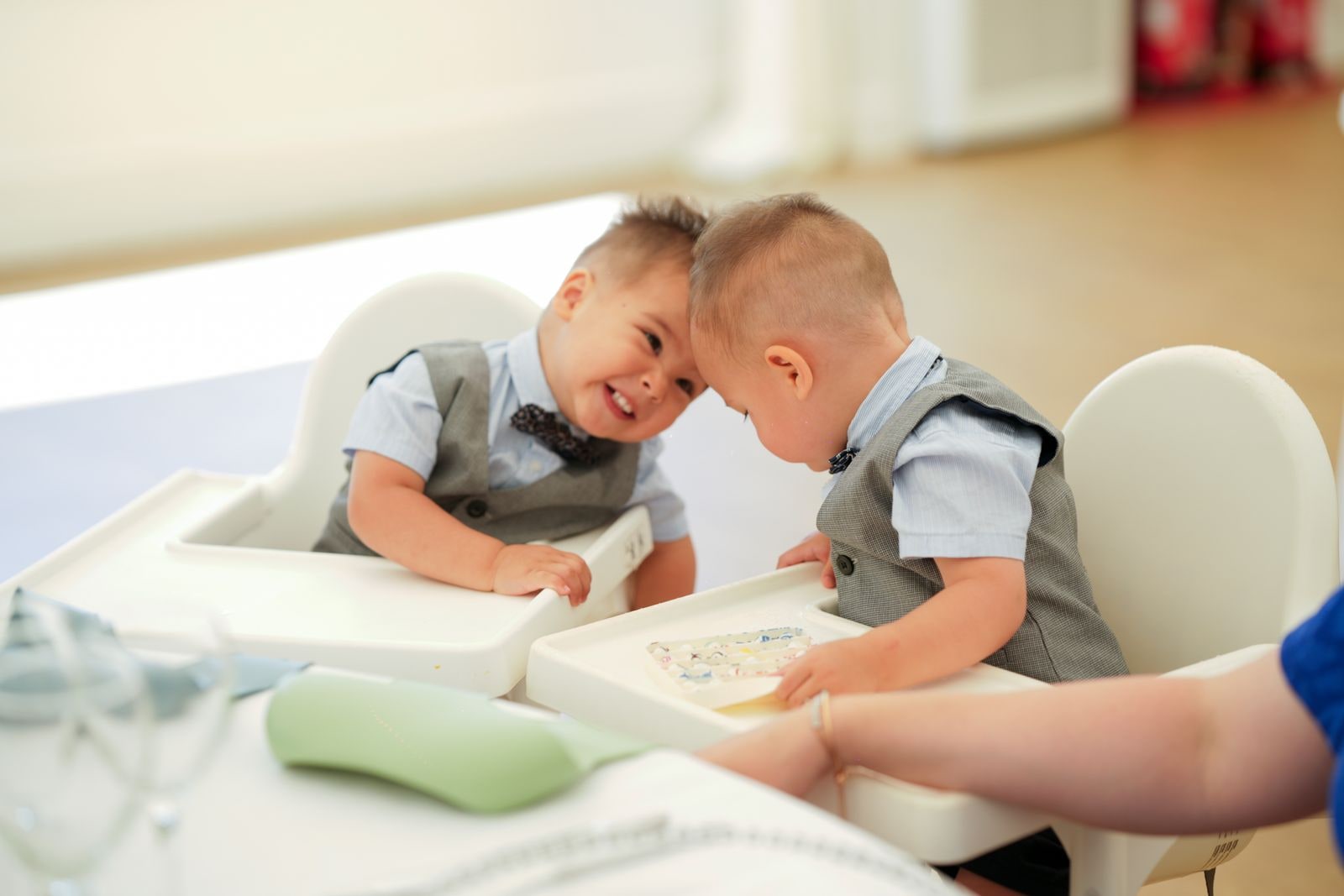 Two small boys in bow ties and waistcoats leaning their heads together in high chairs