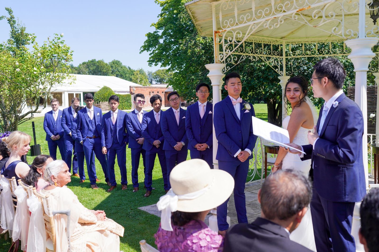 Groomsmen in blue suits lined up beside the bandstand as the celebrant reads to the couple