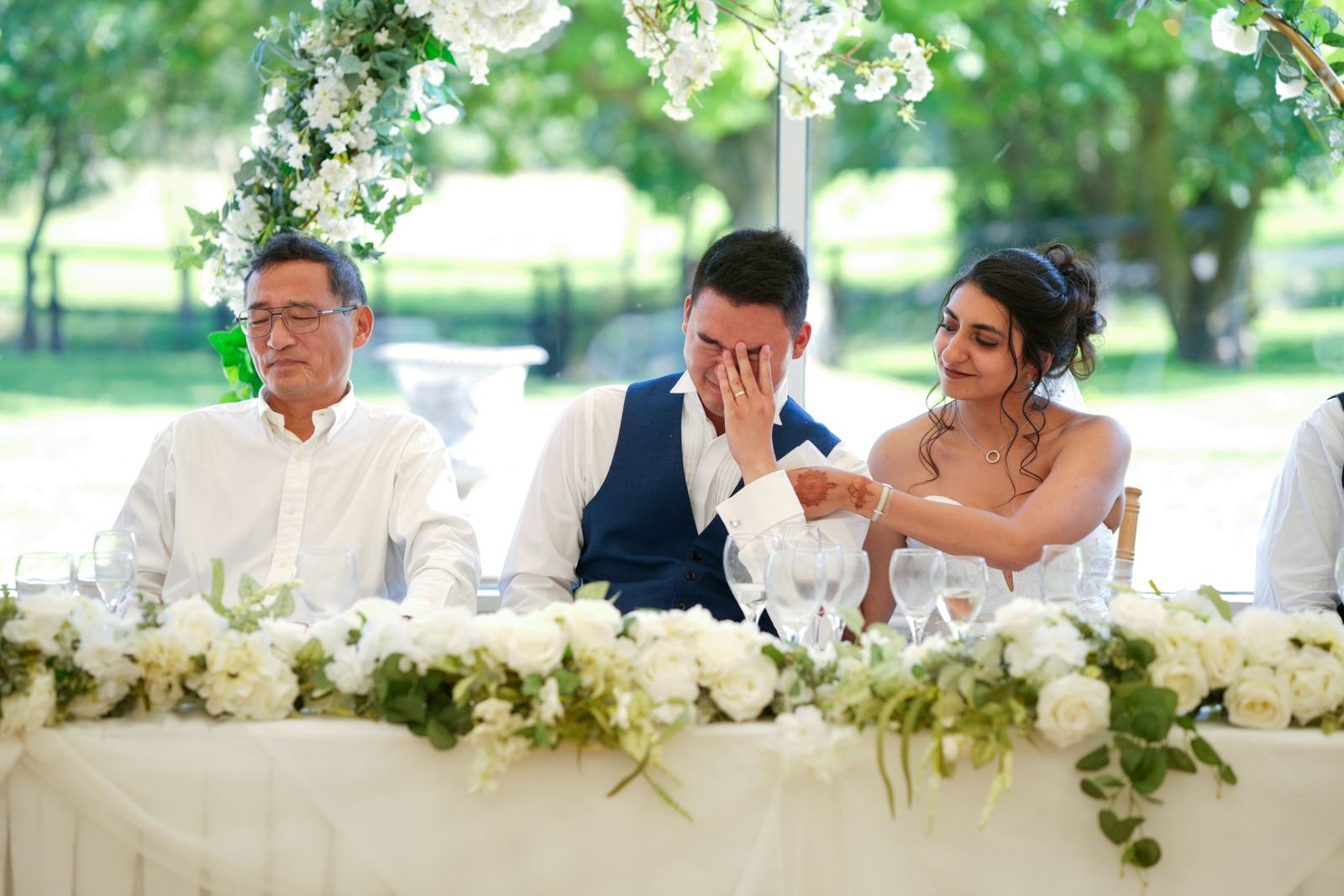 Groom wiping his eyes at the top table, the bride's hand on his arm, white flowers along the front