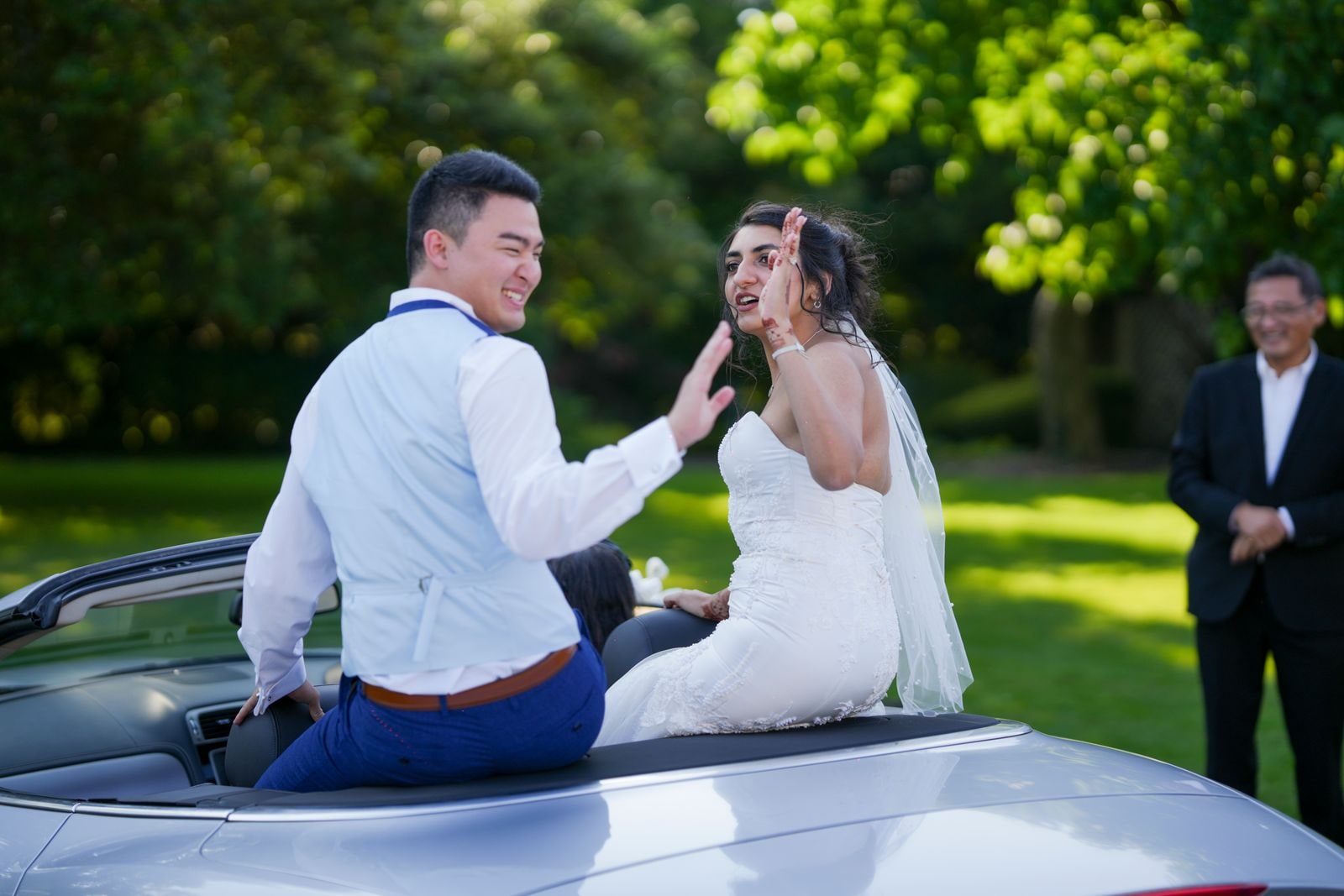 Bride and groom sitting up on the back of an open-top car, laughing together