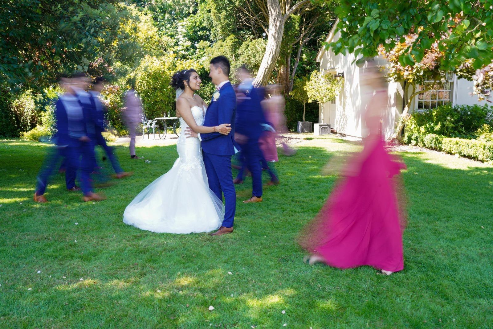 Bride and groom still at the centre as guests move around them in a blur of blue and pink
