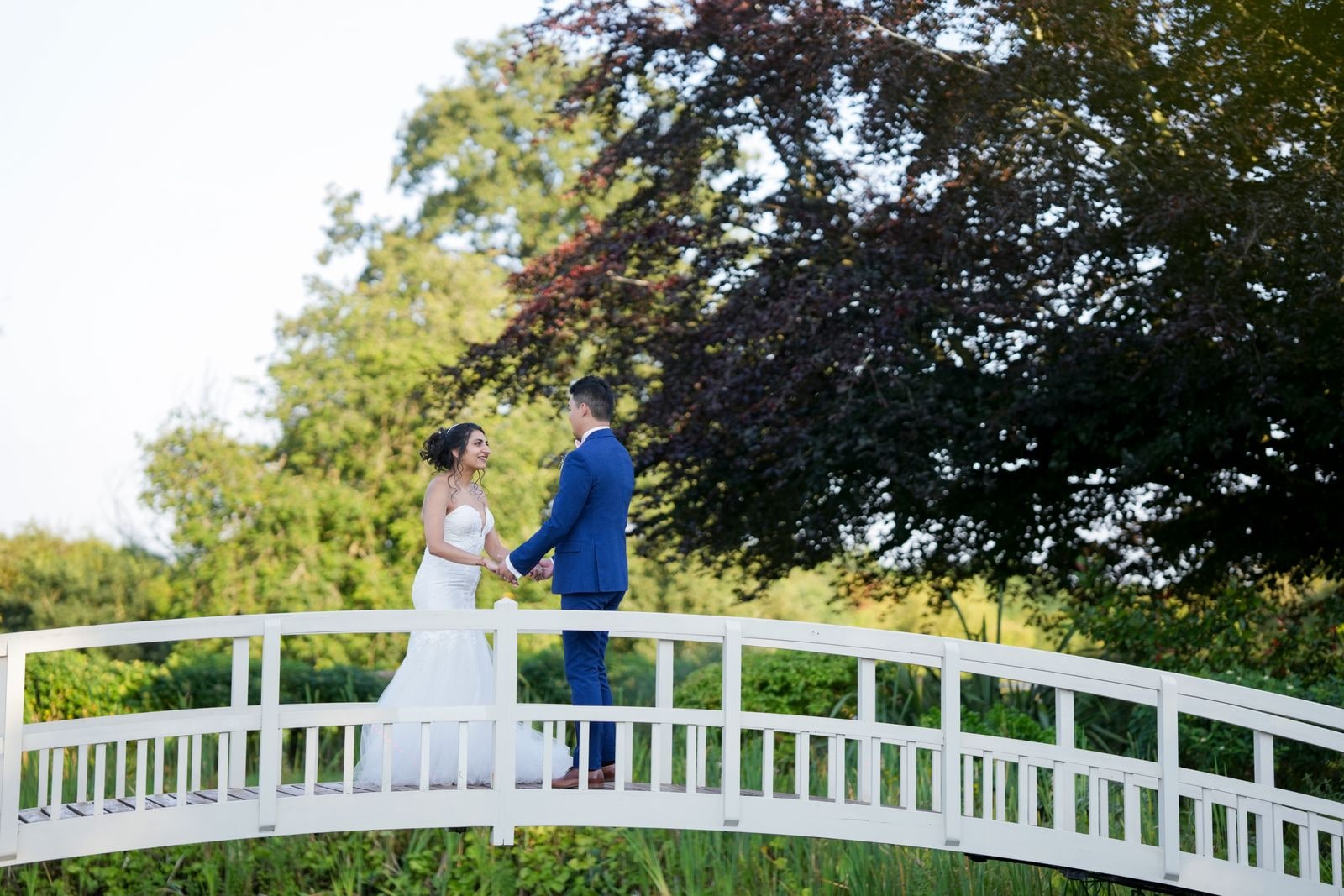 Bride and groom holding hands on the white footbridge, framed by trees