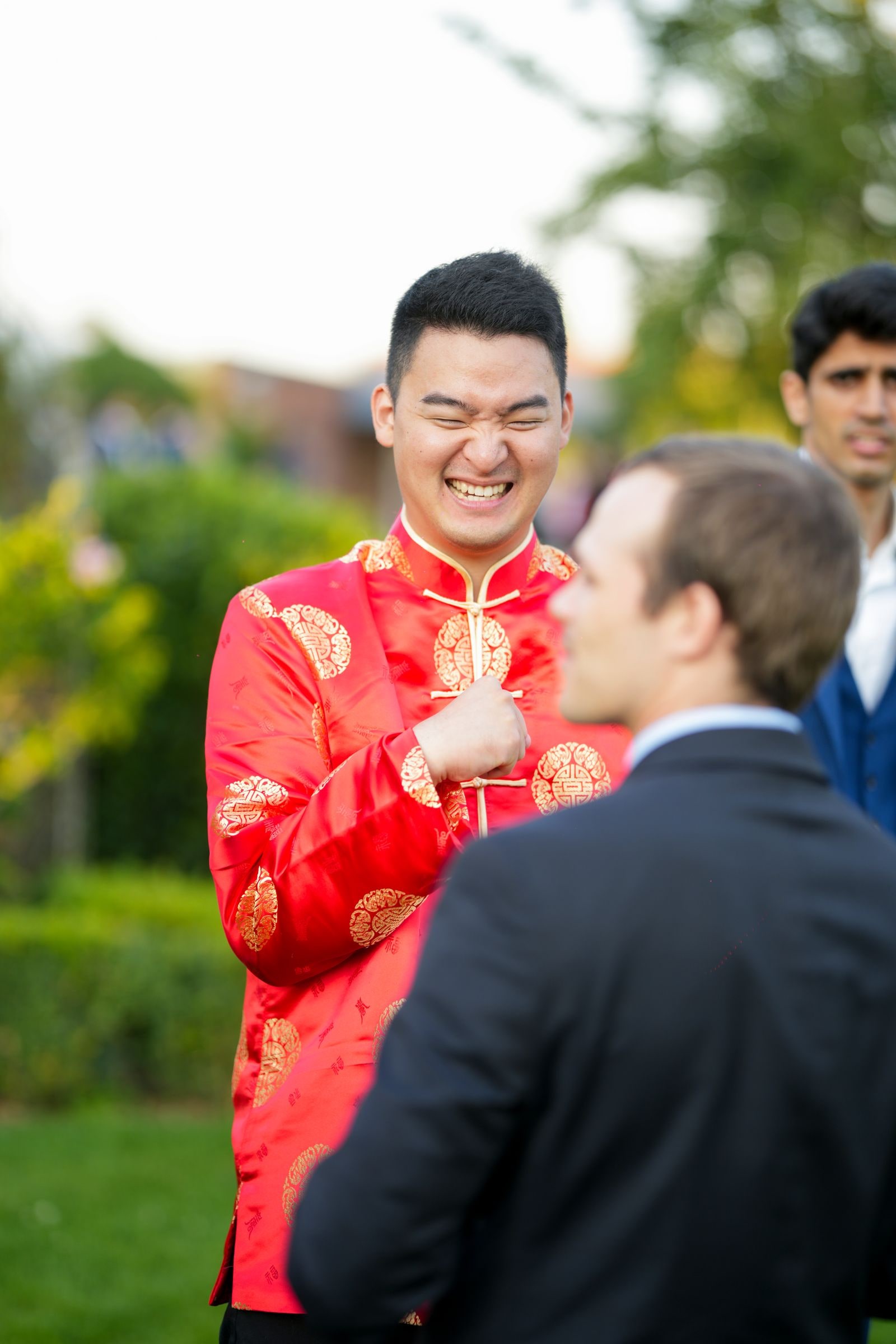 Groom in a red and gold Chinese jacket laughing with a guest at the reception