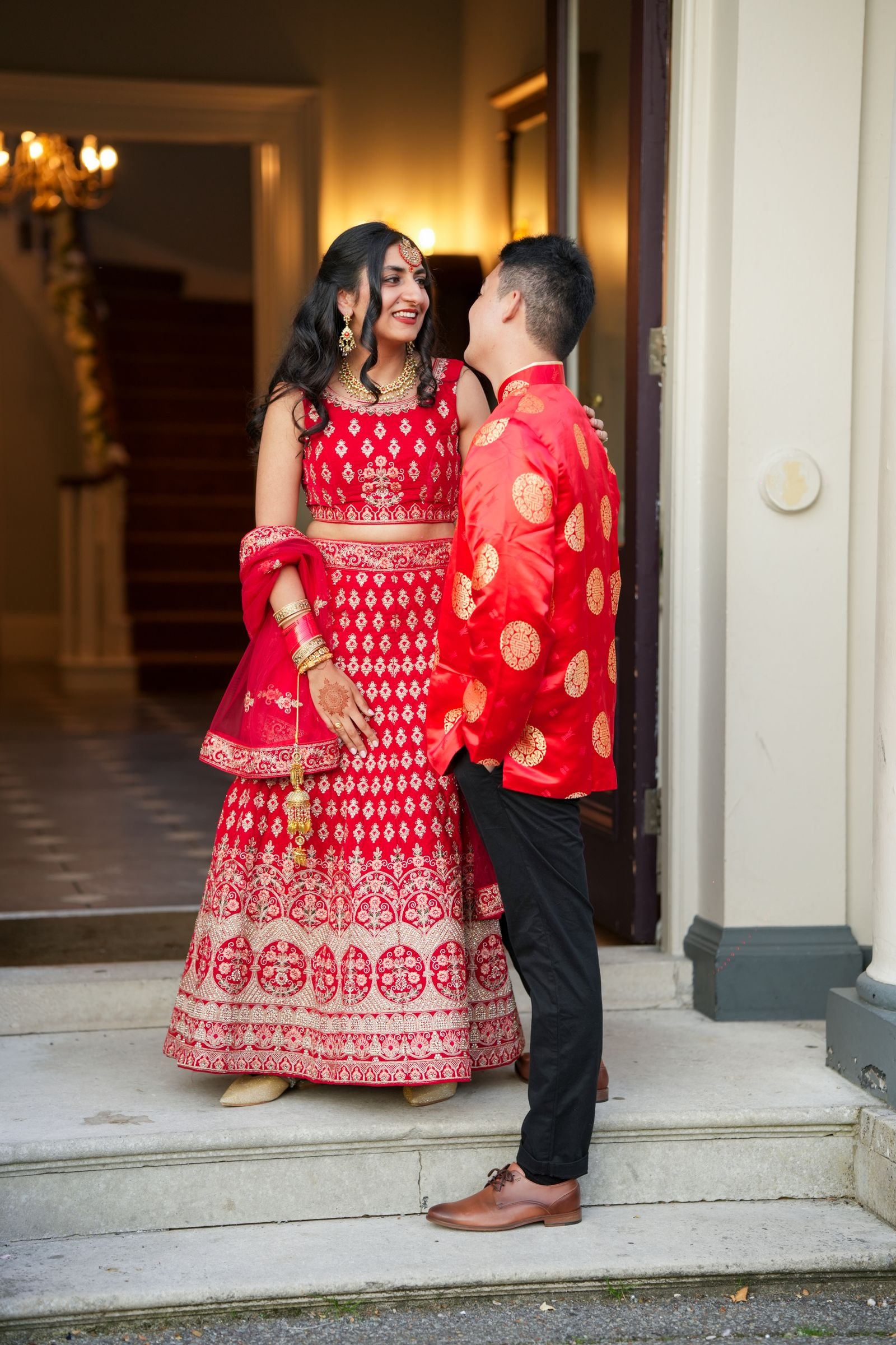 Groom in a red jacket leaning to his bride on the front steps of the house