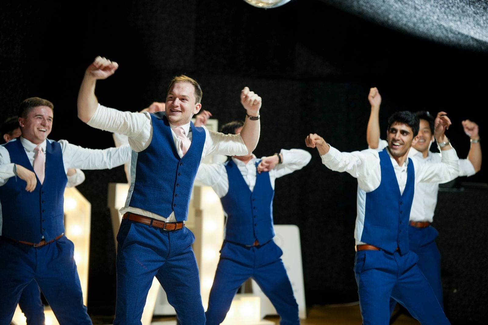 Groomsmen in blue waistcoats mid dance routine, arms raised in a line
