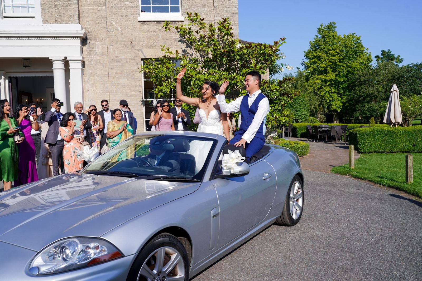 Guests cheering from the terrace as the couple pull away in a silver convertible