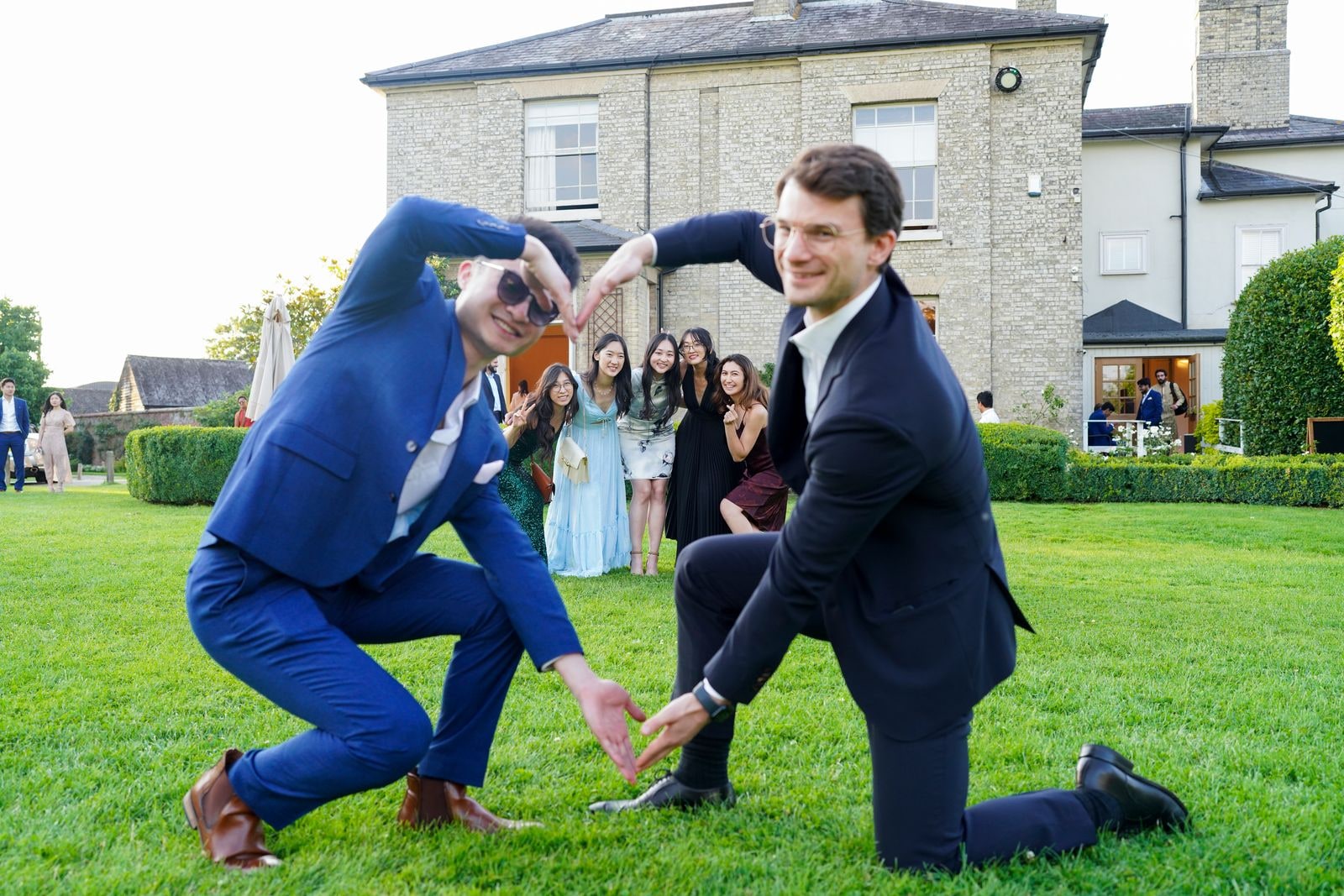Two guests in suits clowning on the lawn, one kneeling, while others watch