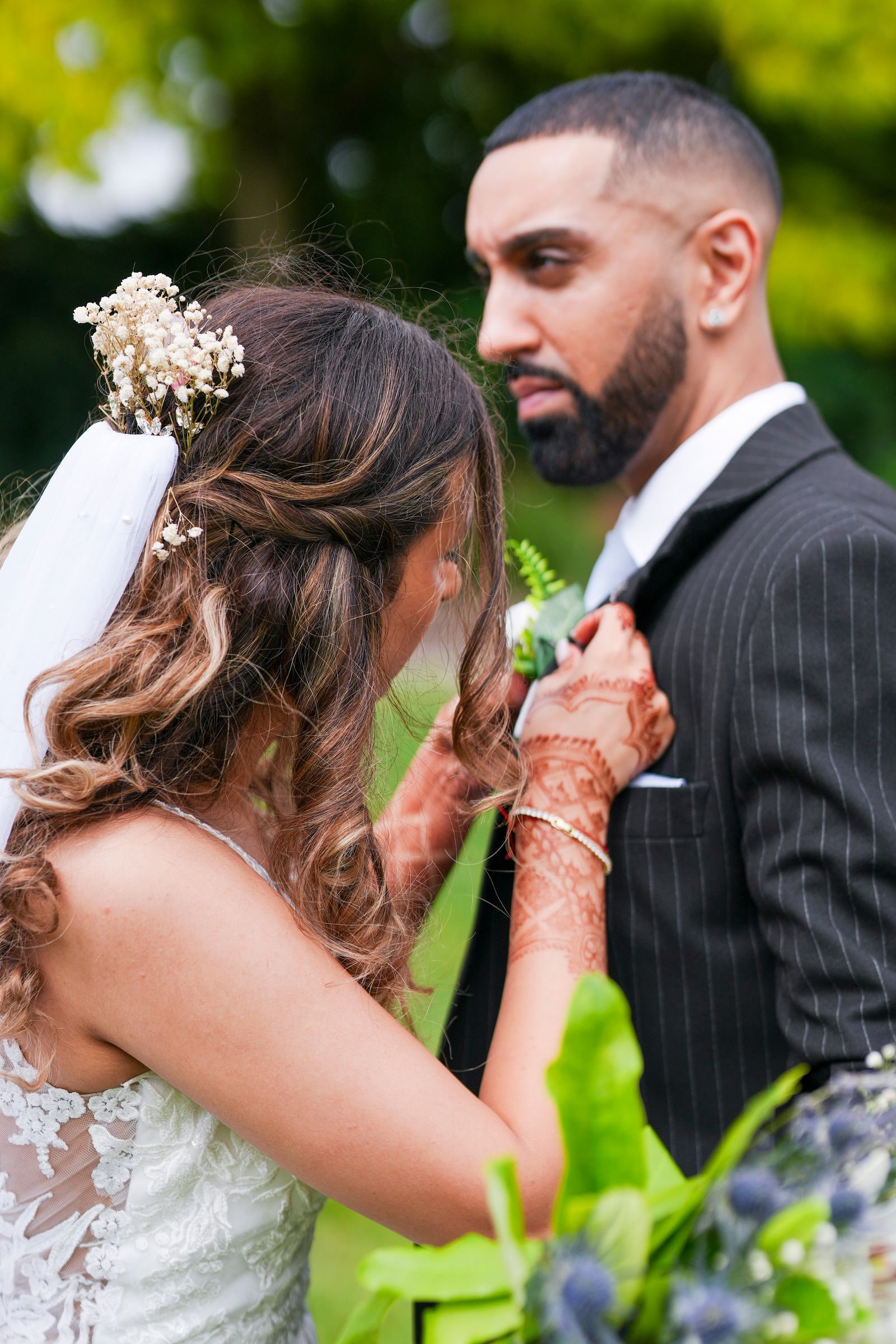 Bride pinning the buttonhole to her groom's pinstriped jacket, her hands hennaed