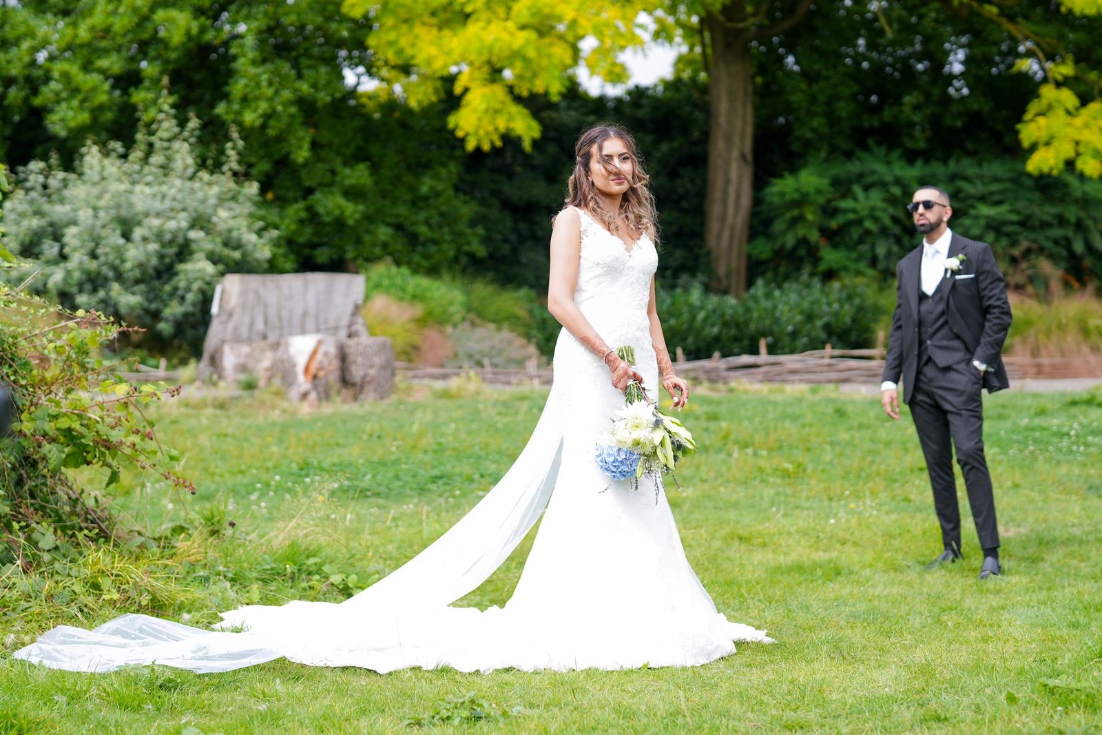 Bride with a blue and white bouquet on the grass, her groom standing apart from her