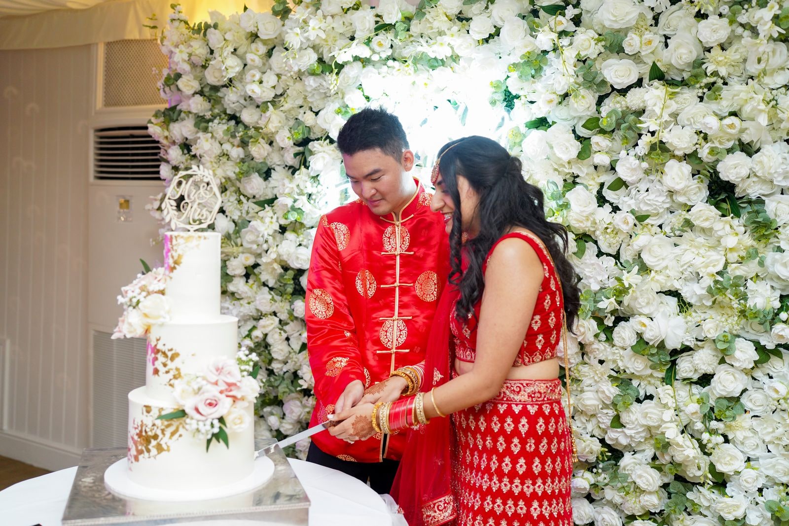 Couple cutting the tiered white cake in front of a wall of white roses