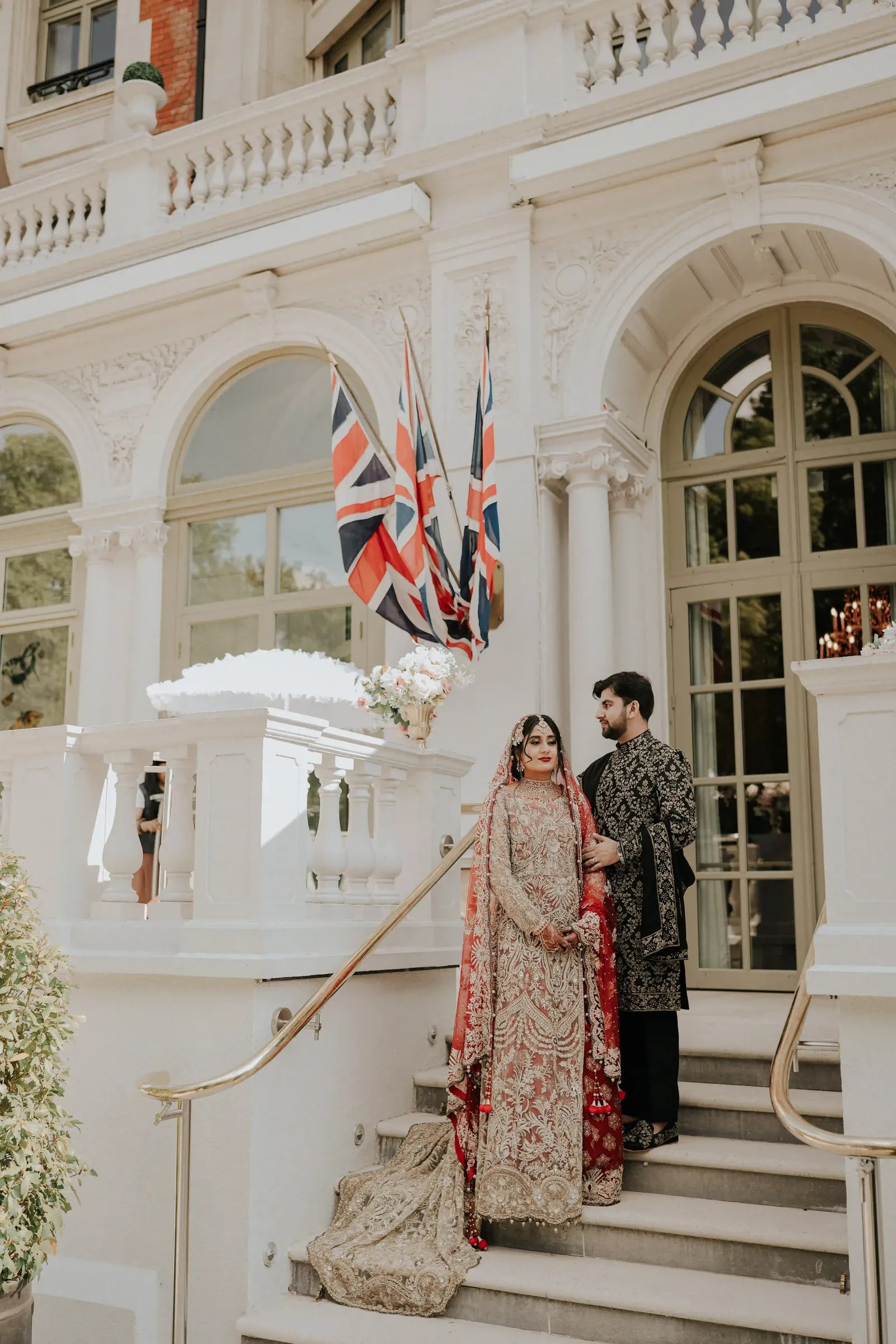 Bride in gold and groom in a dark sherwani on the steps beneath the flags