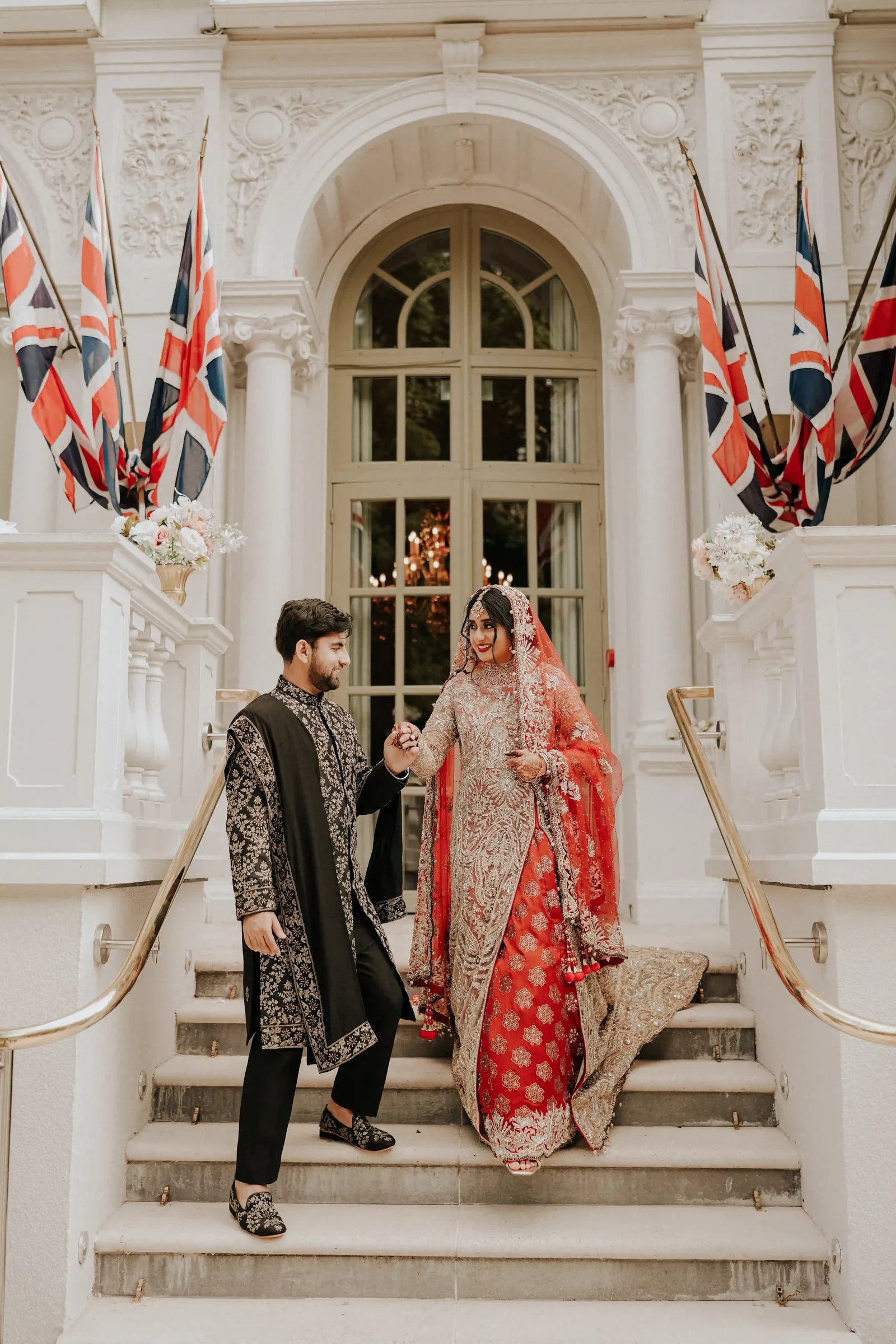 Groom leading his bride down the hotel steps, her red lehenga trailing behind