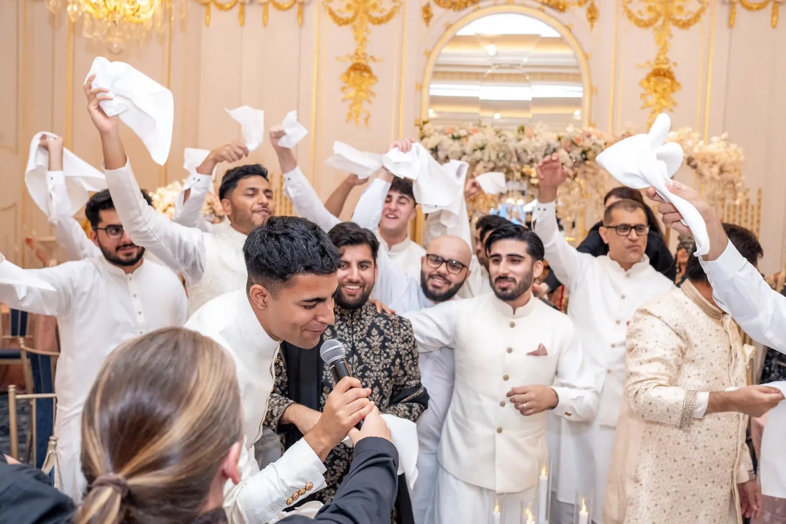 Groom singing into a microphone as his friends whirl napkins above him