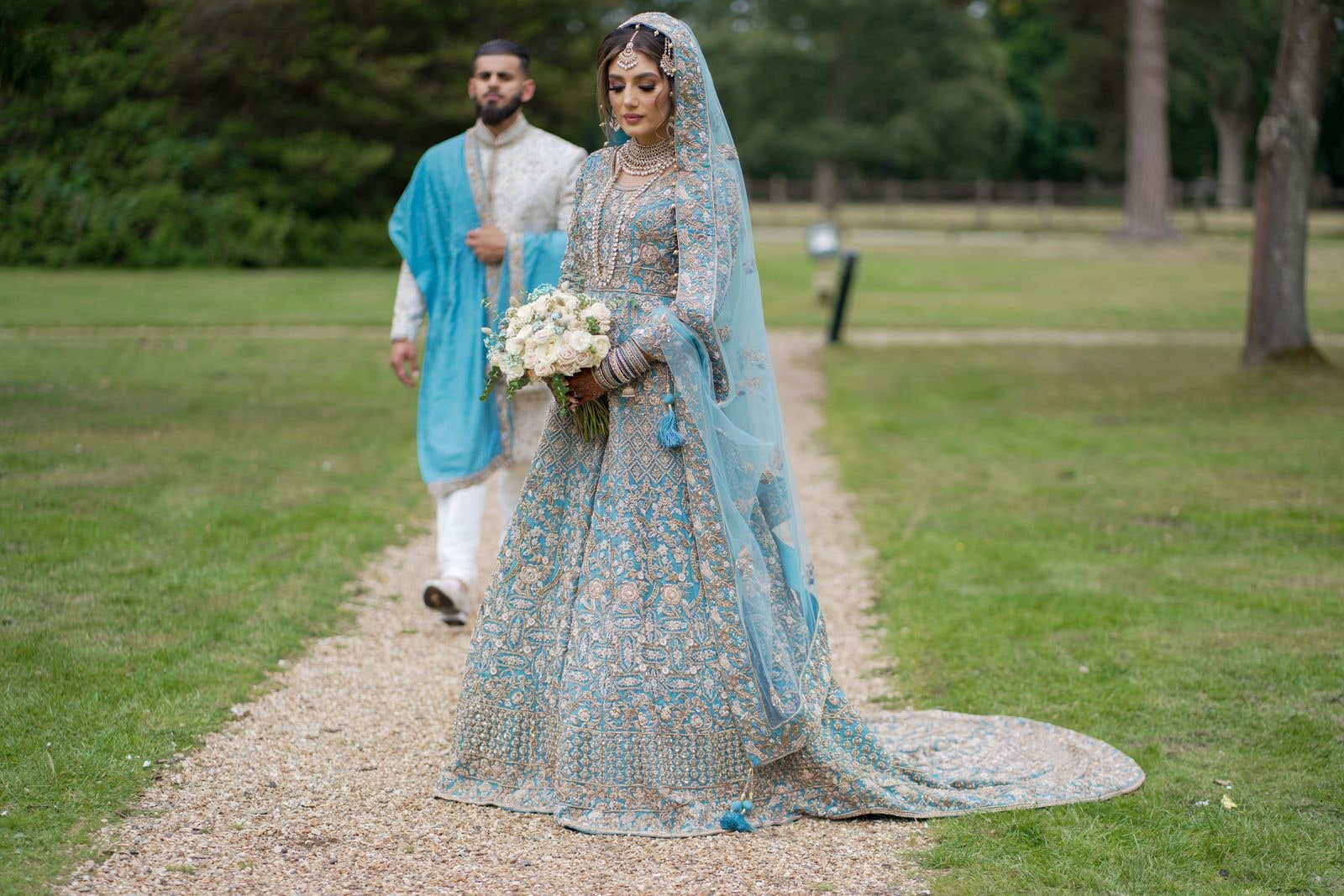Bride in a pale blue and gold lehenga walking a gravel path with her groom, her train trailing behind