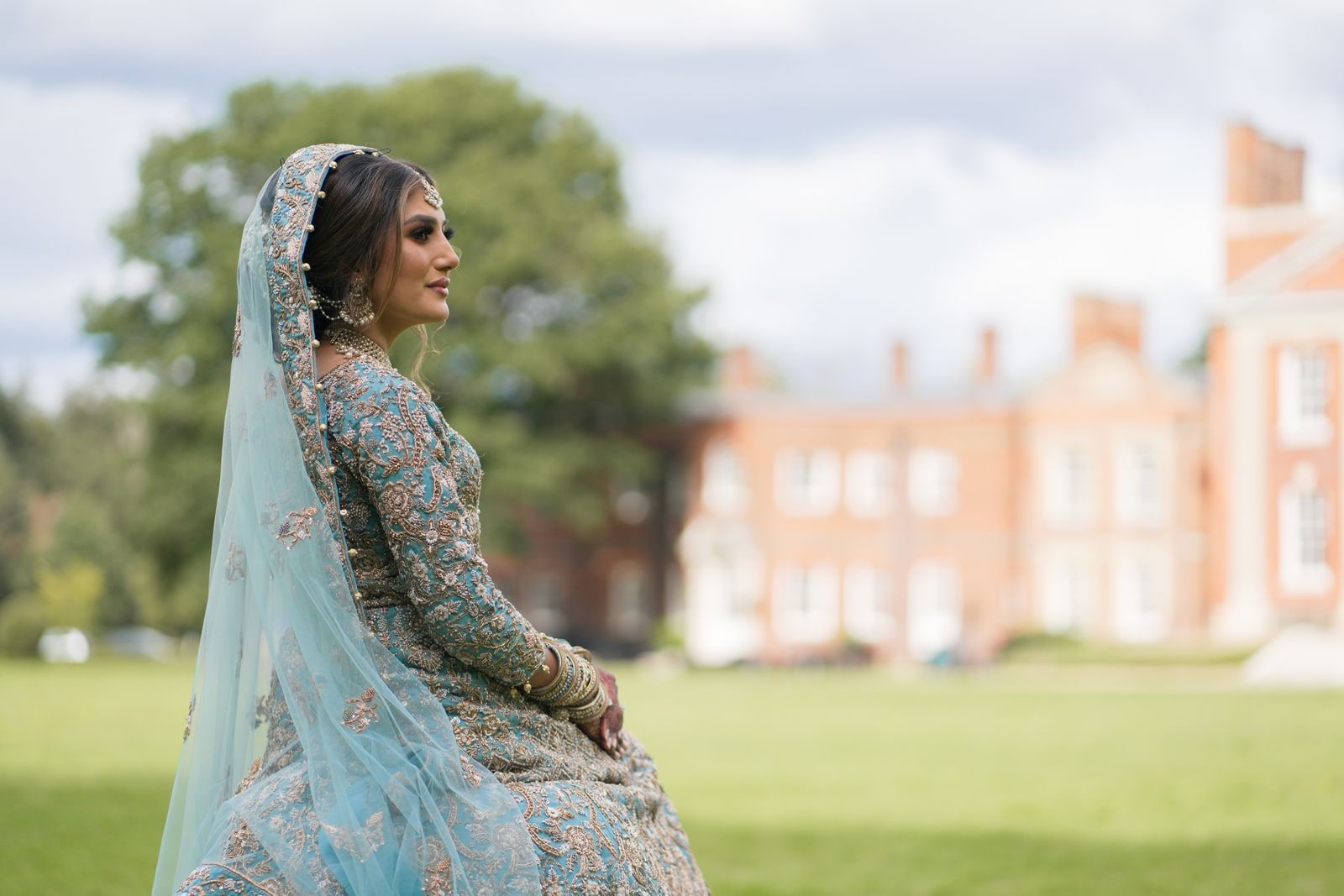 Bride seated on the lawn in a pale blue lehenga, the red brick house behind her