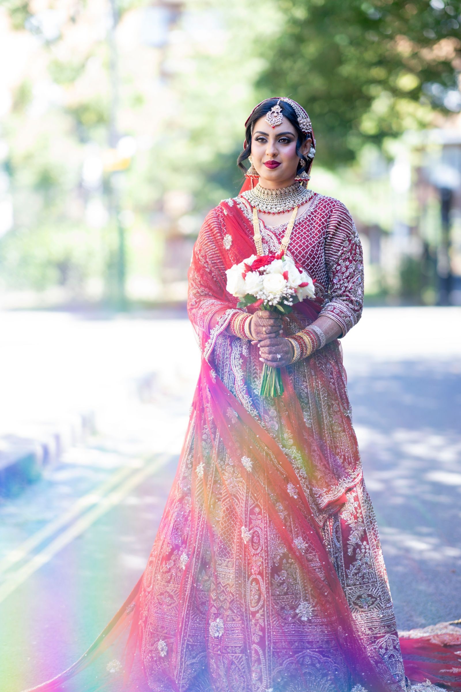 Bride in a red and gold lehenga on a sunlit street, a flare of colour across the frame