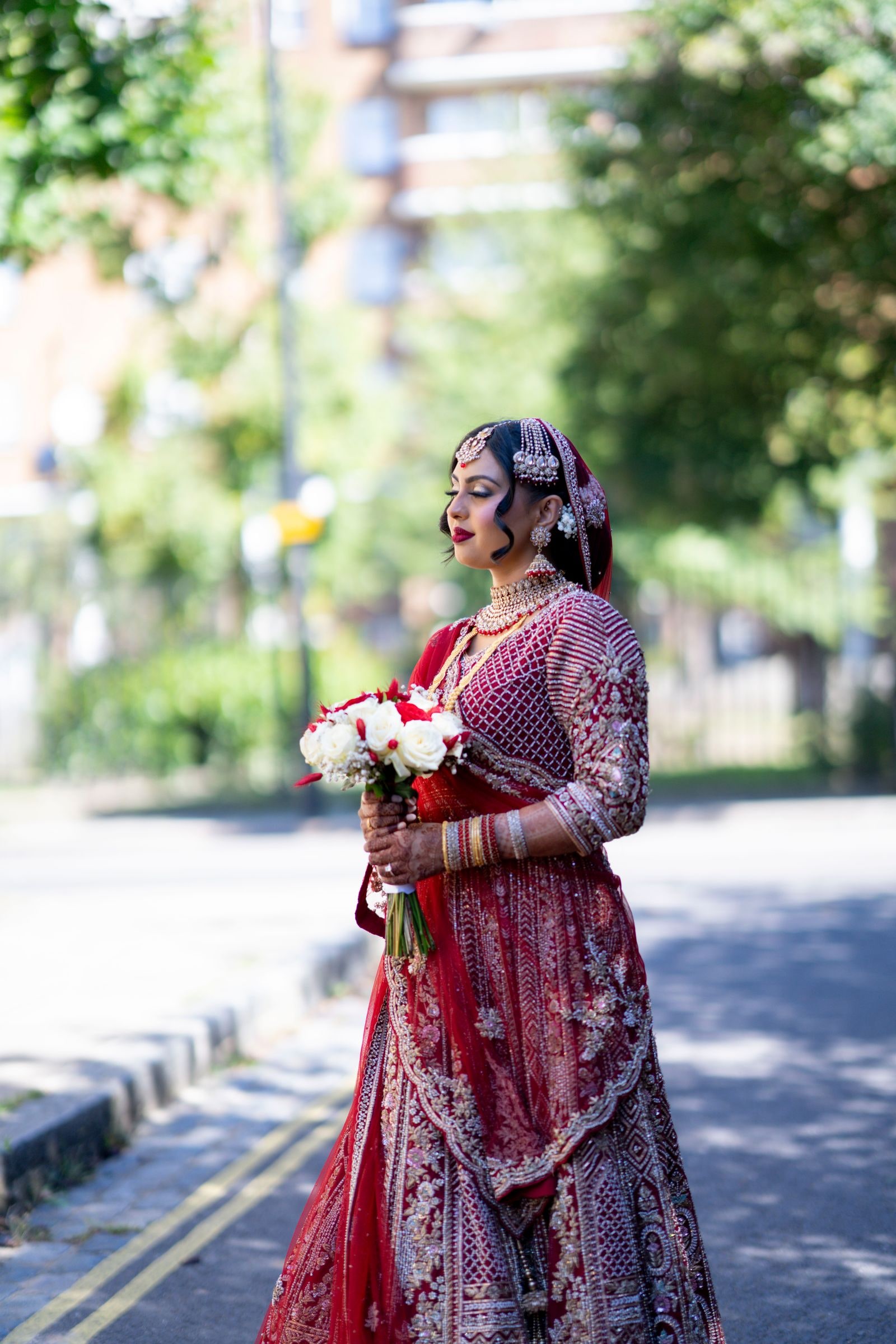 Bride with her bouquet standing at the kerb in a red and gold lehenga